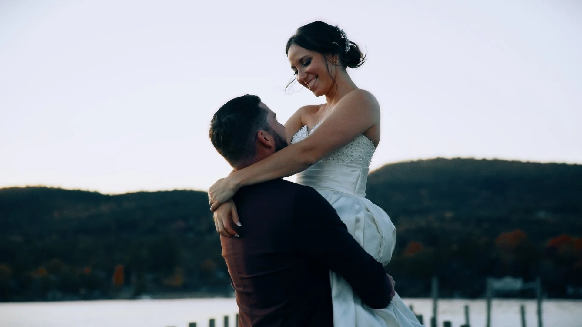 A smiling woman in a wedding dress is lifted by a man in a dark suit at a lakeside during sunset, with hills in the background.