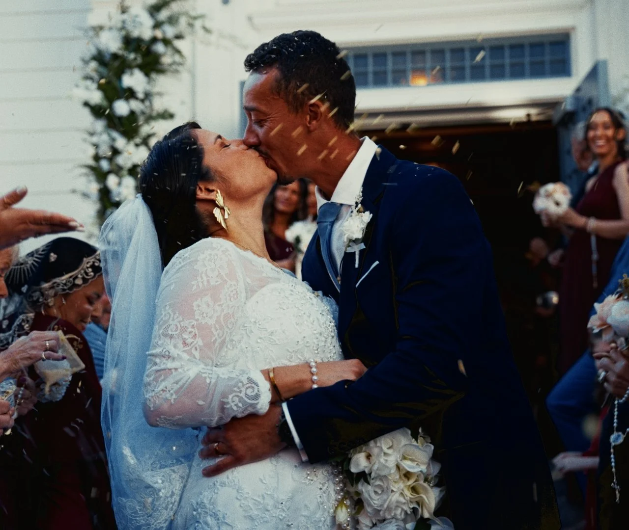 A newlywed couple sharing a kiss at their wedding with guests around them, confetti falling, and a floral arch in the background.