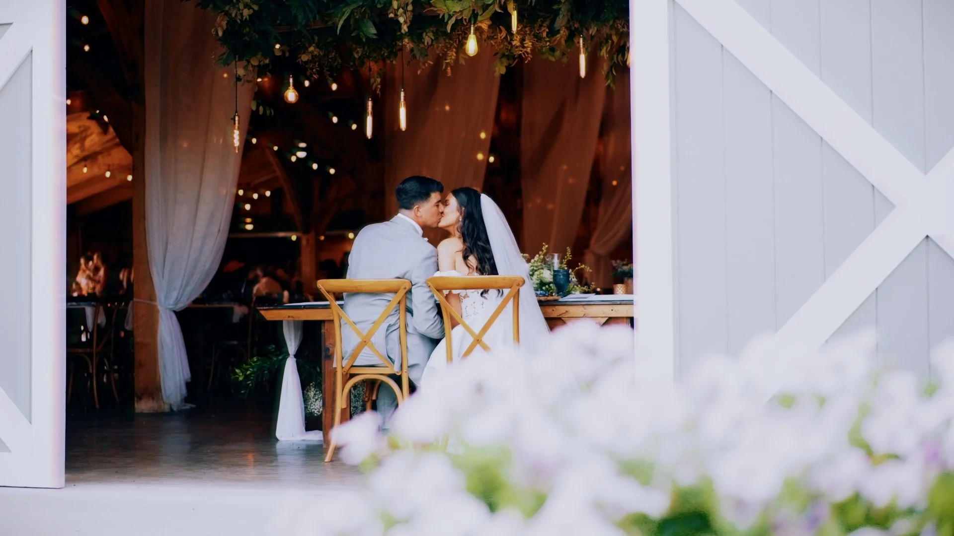 A bride and groom sharing a kiss at their wedding reception, sitting at a wooden table in a rustic setting with warm lighting and draped curtains.