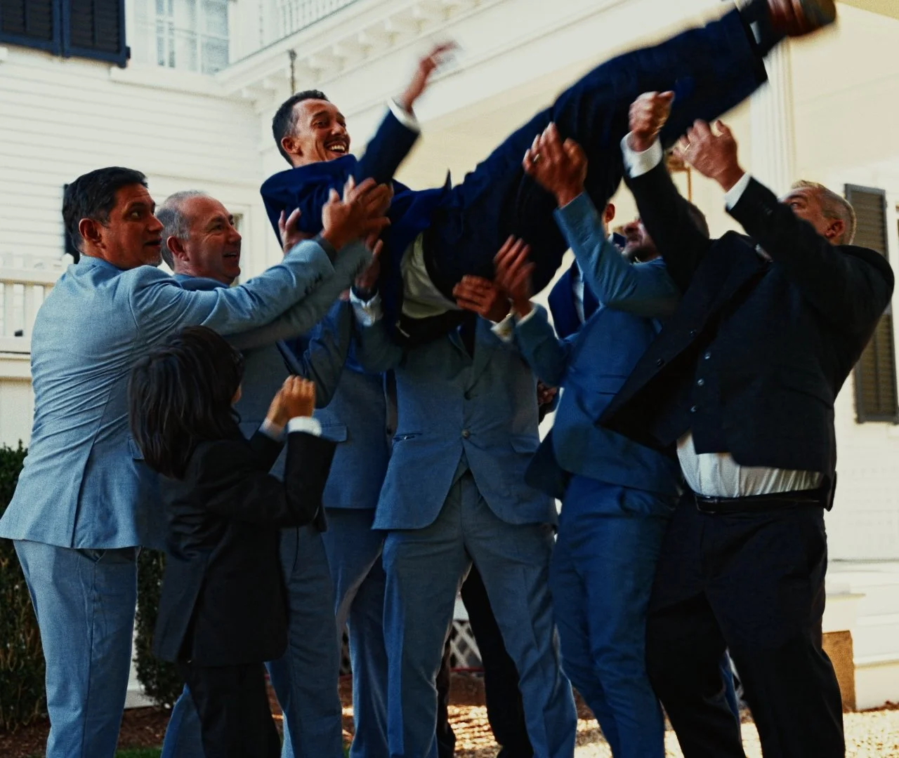Group of people celebrating as they lift a man in a suit into the air in front of a house.