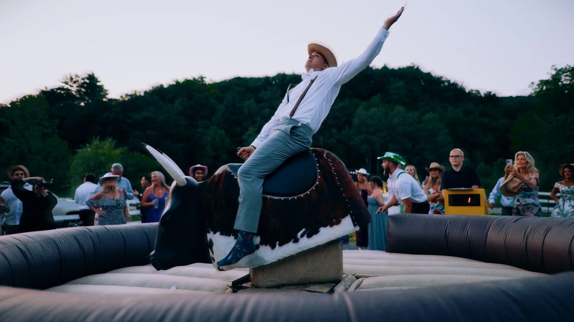 Person dressed in vintage clothing with a straw hat, riding a mechanical cow during a rodeo event, while several people watch and take photos in the background.