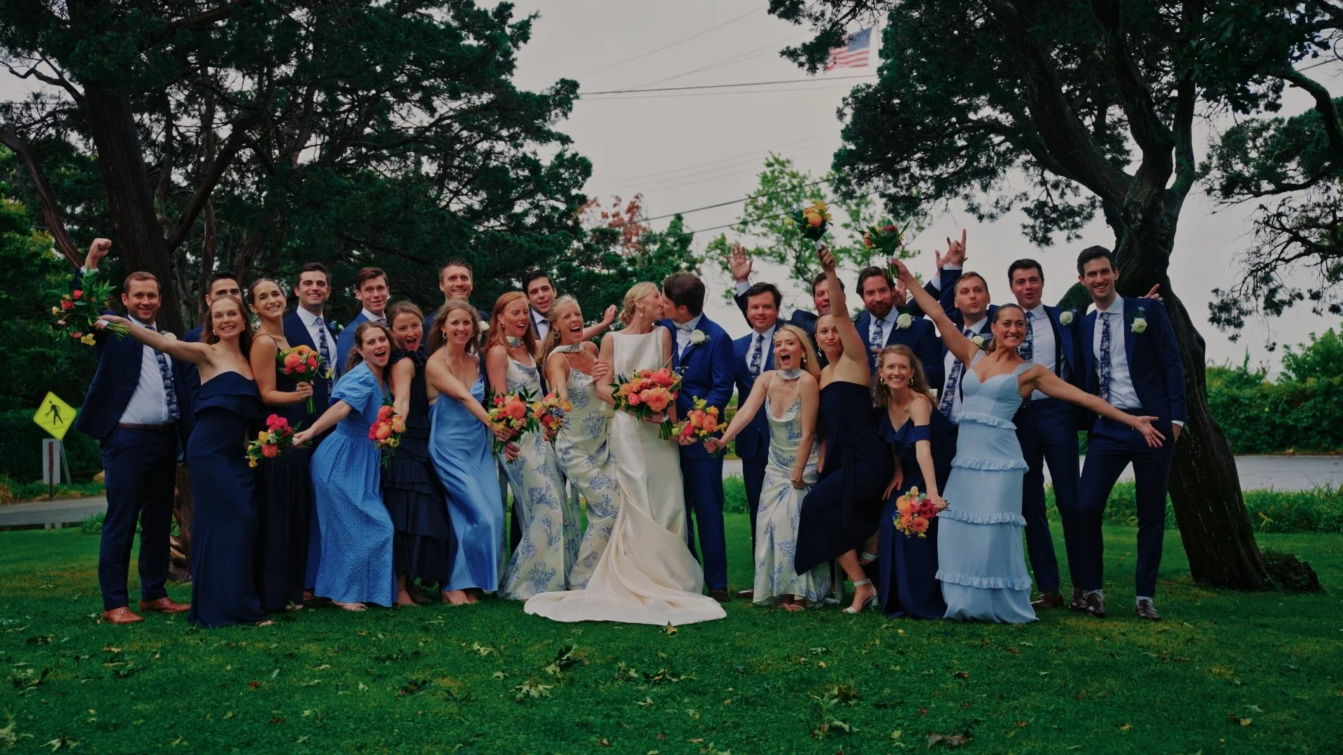 A large group of smiling people at a wedding, with some holding bouquets, standing outdoors on a grassy lawn near trees, celebrating happily.