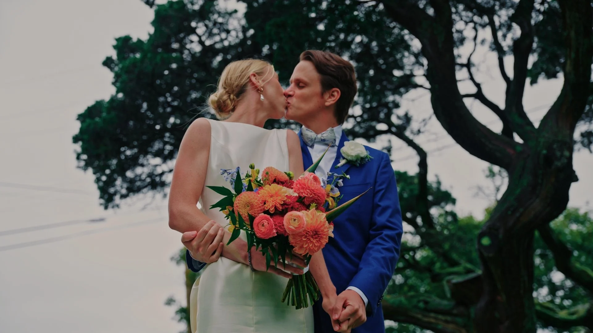 A couple in wedding attire sharing a kiss outdoors, with the woman holding a bouquet of colorful flowers and the man dressed in a blue suit with a bow tie, standing in front of a large tree.