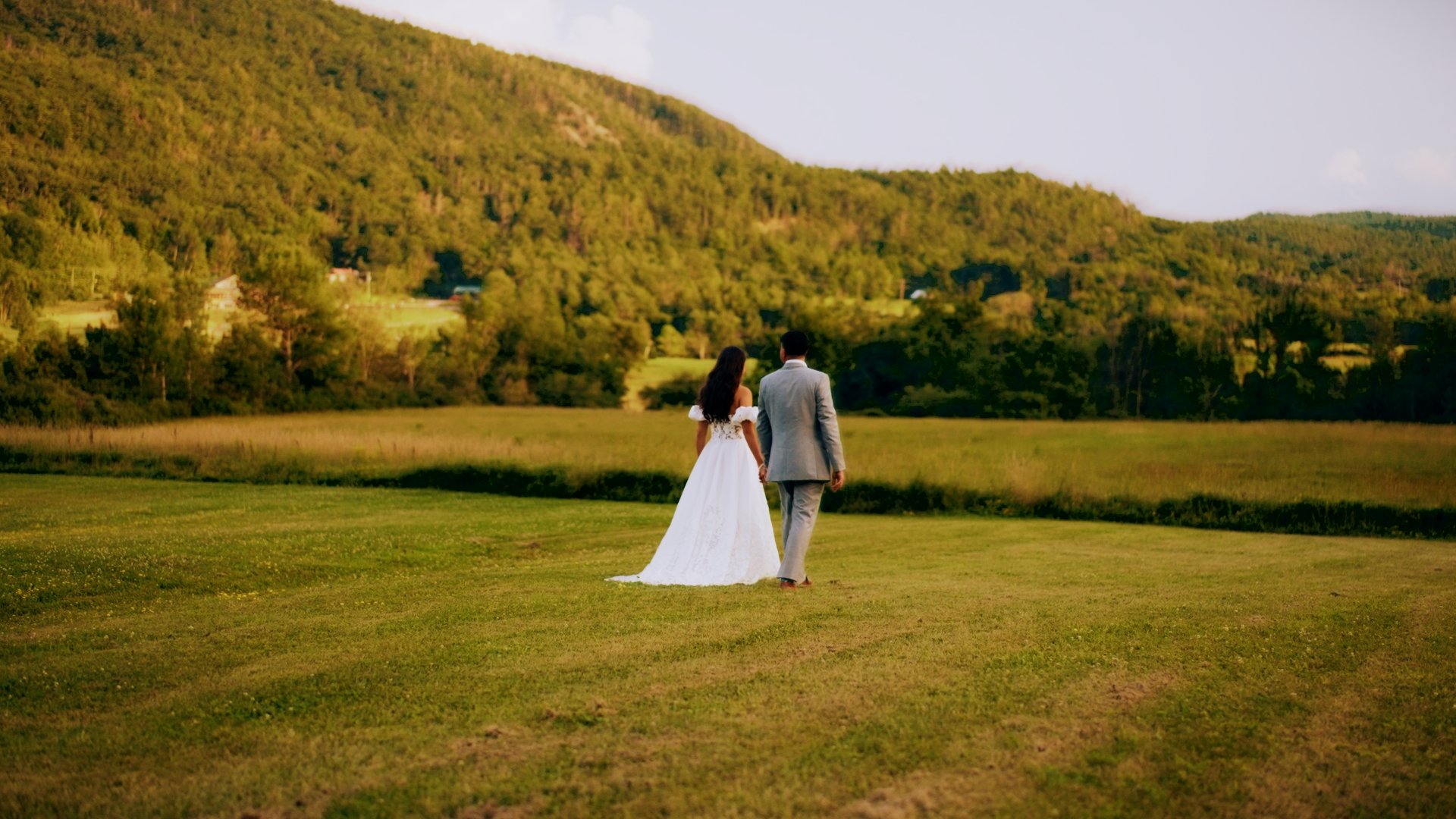 A bride and groom walking hand in hand across a grassy field surrounded by trees and hills during sunset.