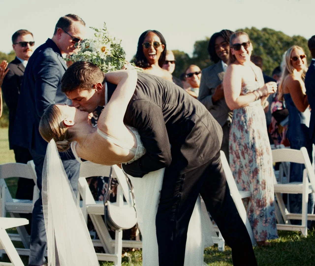 A couple kissing at their outdoor wedding ceremony, surrounded by friends and family.