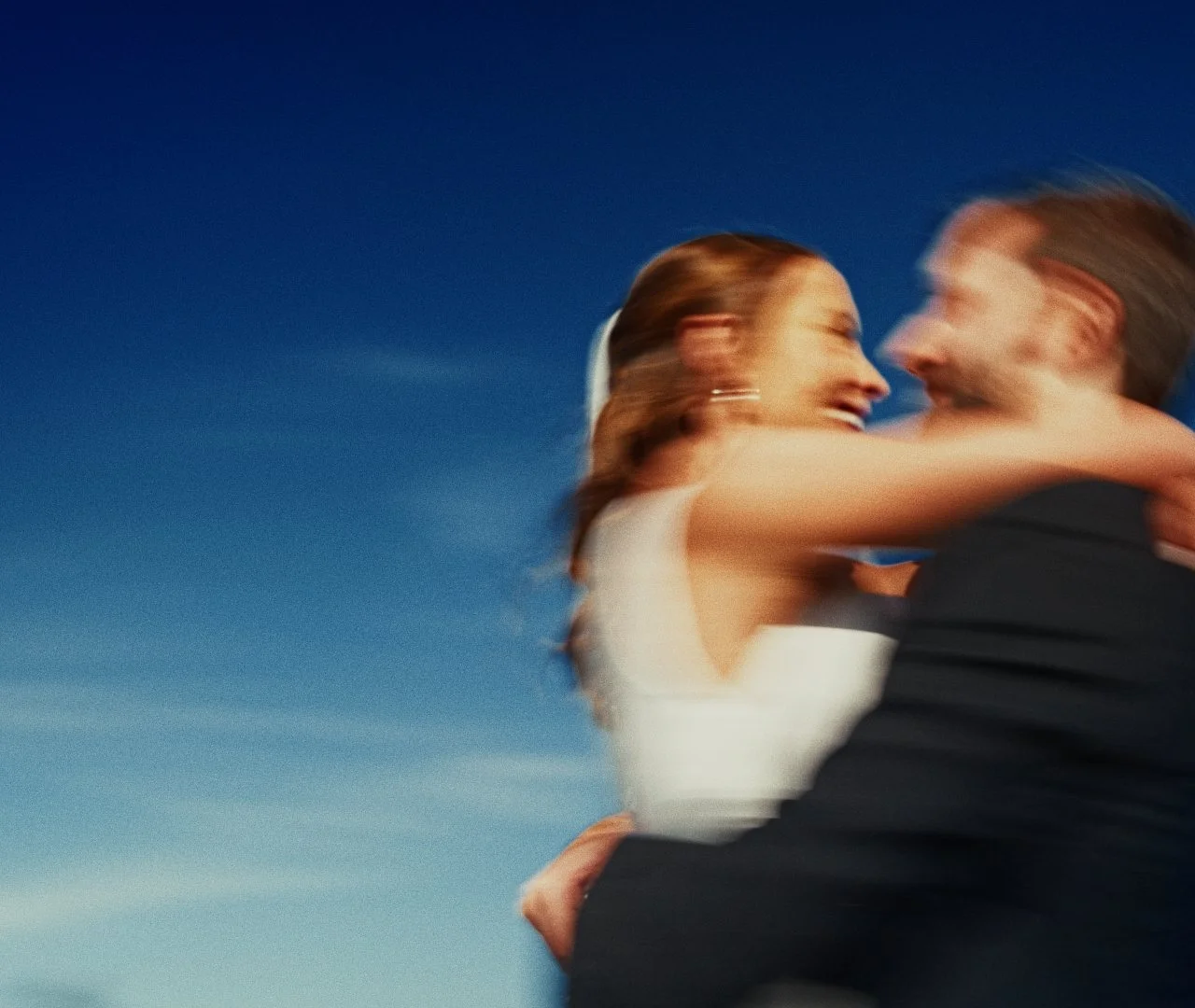 A blurry image of a woman and a man embracing and smiling, set against a blue sky background.