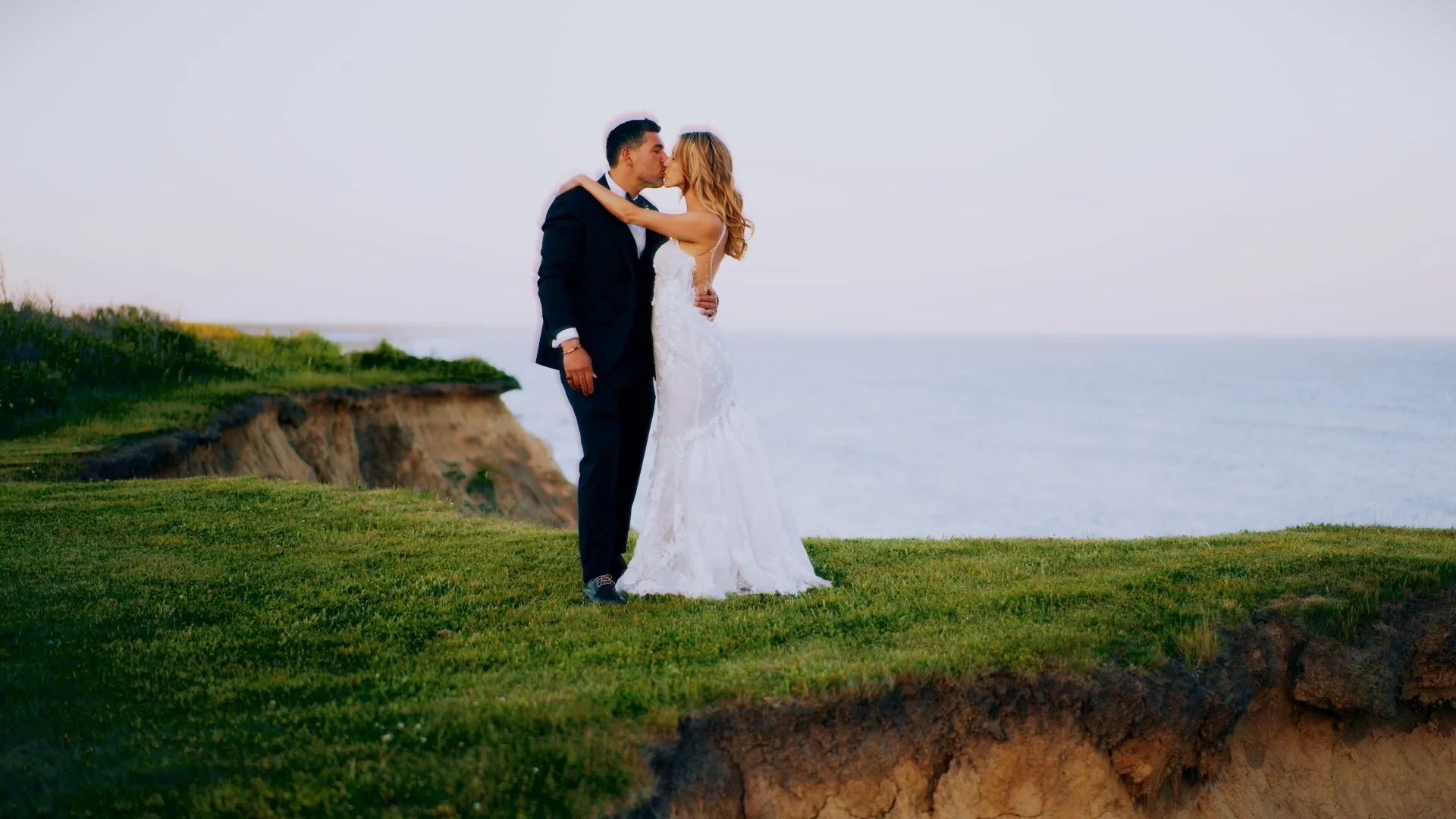 A newlywed couple kissing on a grassy cliffside overlooking the ocean during sunset.