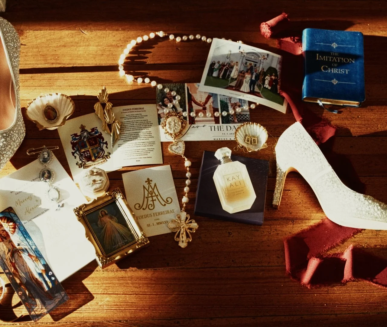 Collection of religious and ceremonial items on a wooden surface, including a Bible titled 'The Imitation of Christ,' religious postcards, jewelry, a framed picture of Jesus, a perfume bottle labeled 'Kay Ali,' a sparkling high-heeled shoe, and vario