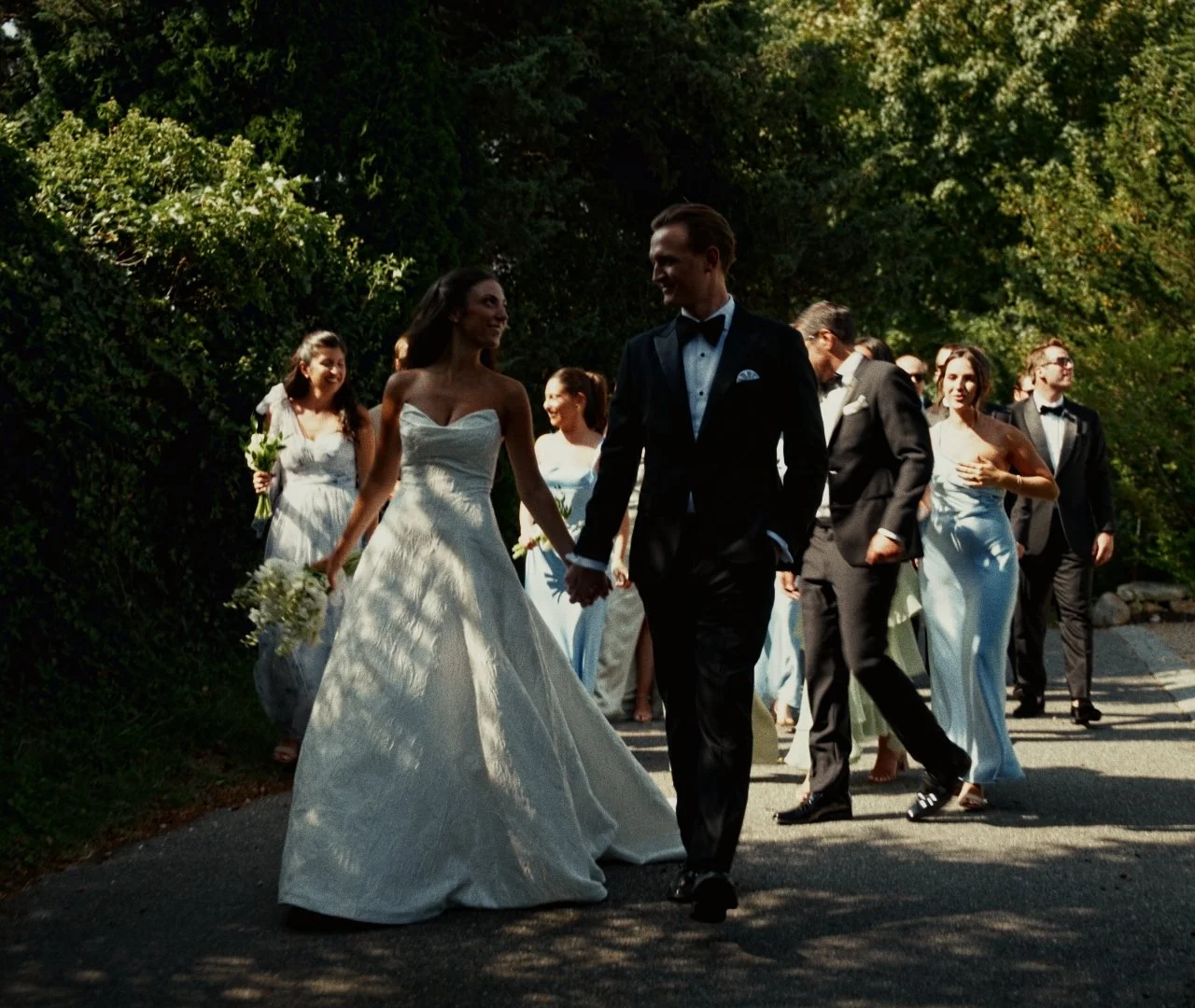 Bride and groom holding hands walking outdoors with wedding party, women in dresses and men in suits, greenery background.
