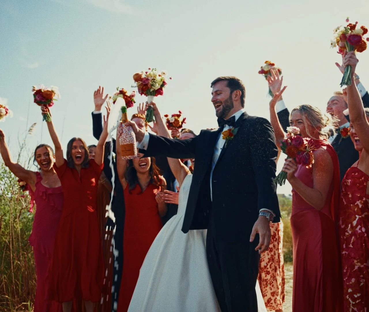 A wedding celebration outdoors with a bride and groom in the center, surrounded by guests in red dresses holding bouquets, cheering and celebrating, with a man pouring champagne.