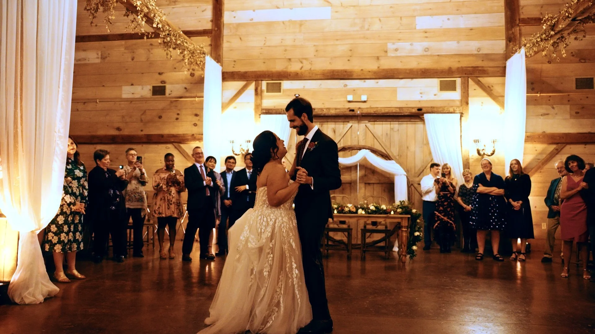 Couple dancing at wedding reception in rustic barn with wood walls, surrounded by guests watching and taking photos.