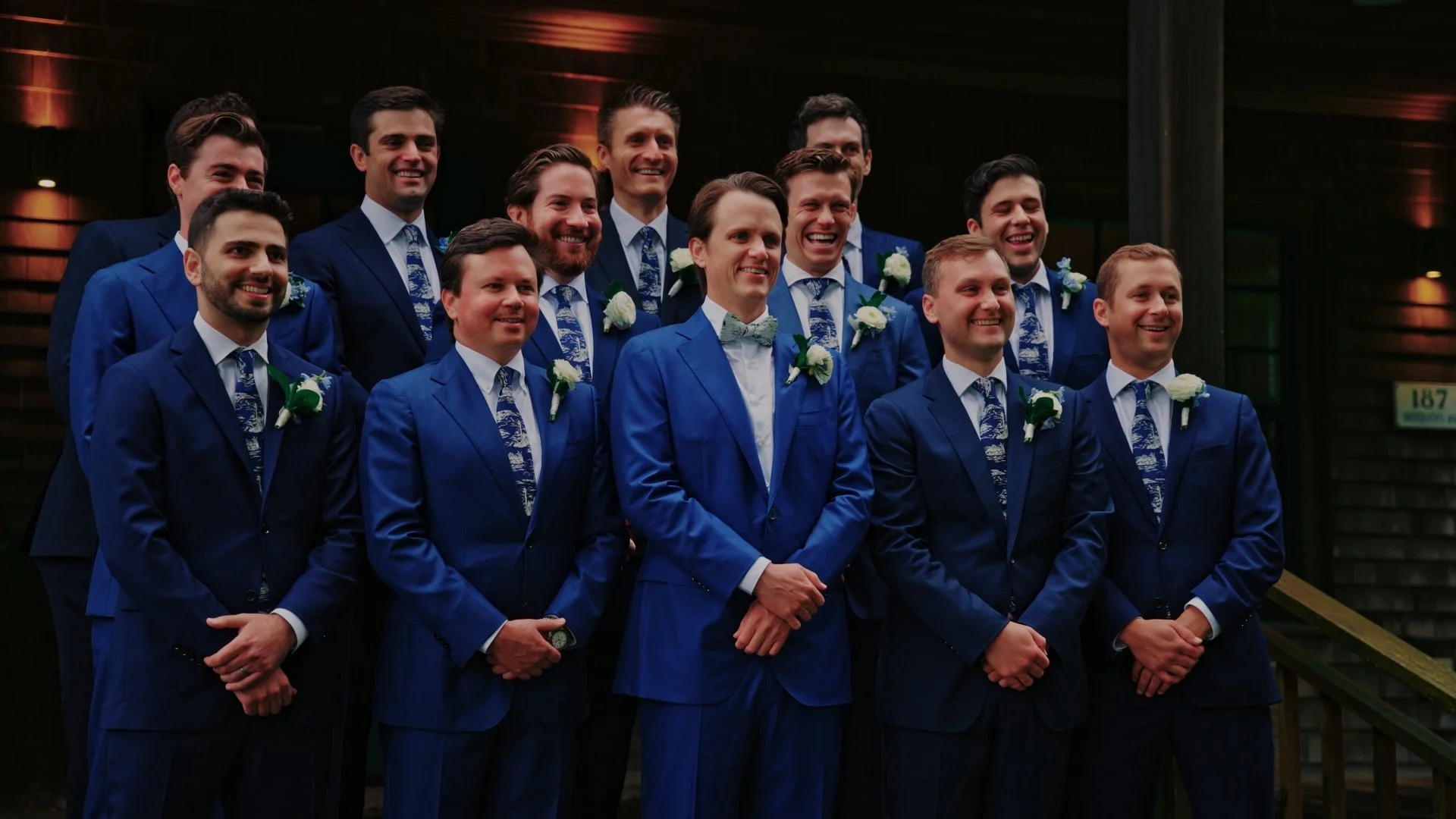 Group of groomsmen and the groom at a wedding, all dressed in matching blue suits with white shirts, patterned blue ties, and white boutonnieres, posed outdoors in front of a wooden building at night.