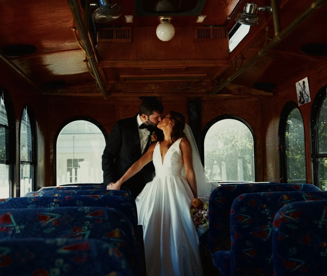 A bride and groom sharing a kiss inside an old-fashioned bus, with wooden interior and patterned blue seats.