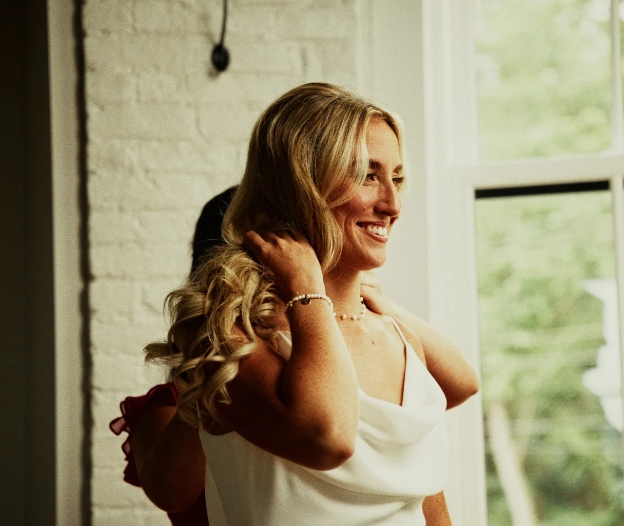 A woman smiling with blonde hair, adjusting her hair, wearing a white dress and jewelry, in front of a window with greenery outside.