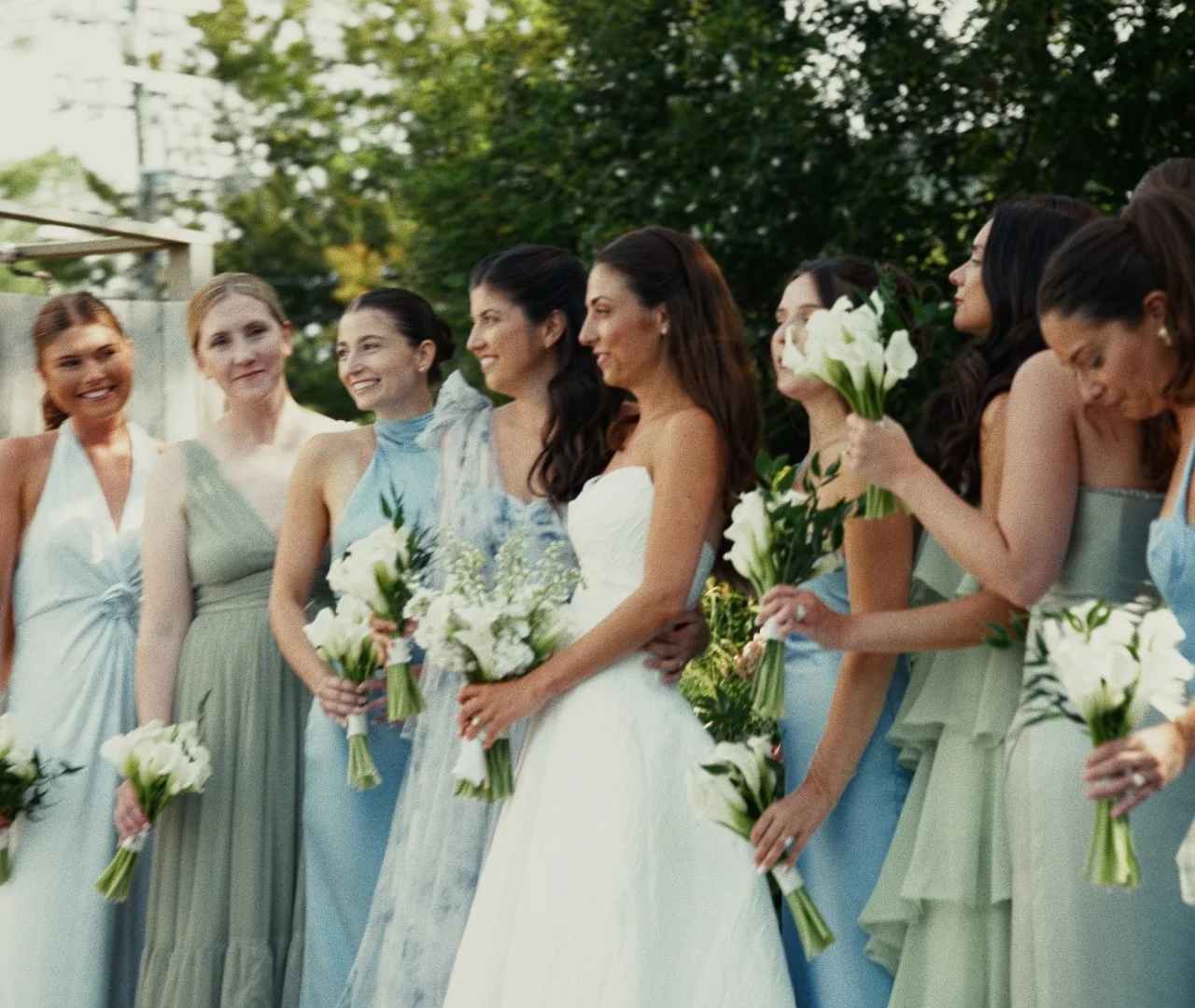 Bride in a white wedding dress and her bridesmaids in pastel dresses holding bouquets of white flowers, outside in a garden setting.