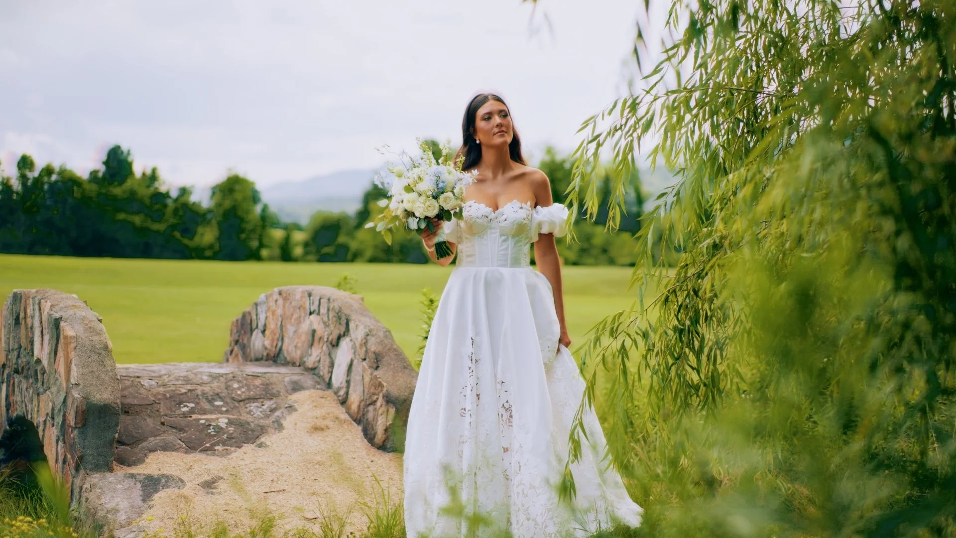 A woman in a white wedding dress holding a bouquet of white and blue flowers outdoors near a stone bridge in a green landscape.