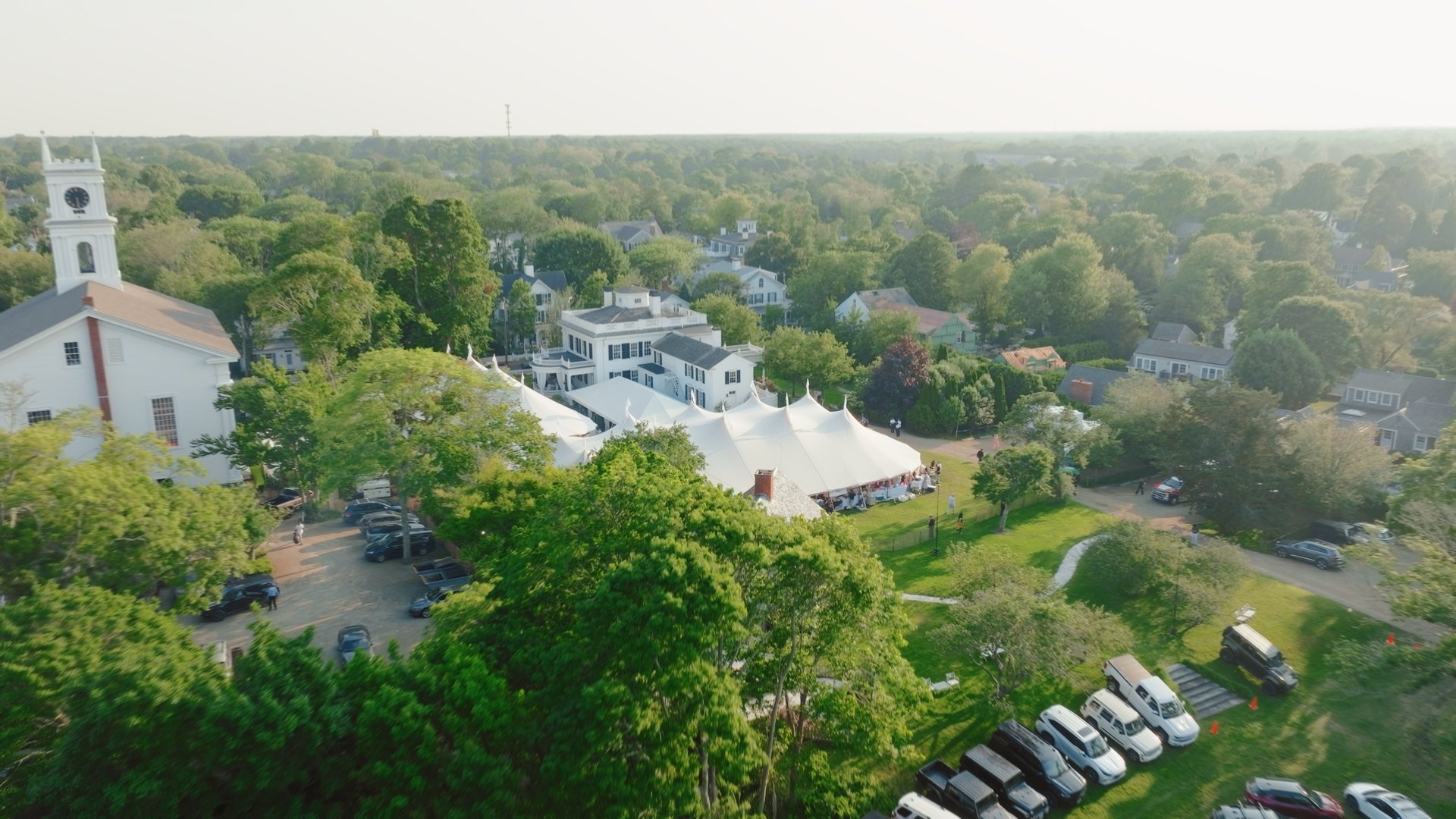 Aerial view of a white tent set up on a grassy area with surrounding trees, adjacent to a white church with a tall clock tower, in a small town with various houses in the background and parked cars in the foreground.