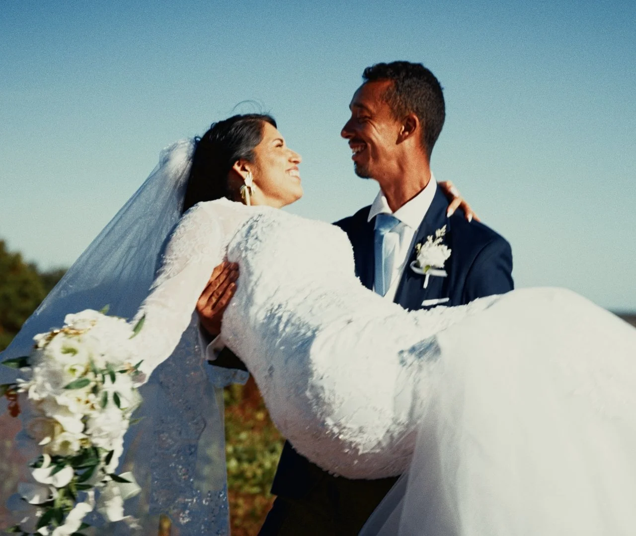 A bride and groom smiling at each other outdoors on their wedding day, with the groom carrying the bride in his arms. The bride wears a white wedding dress and veil, and the groom is in a dark suit with a boutonniere. The bride holds a bouquet of whi