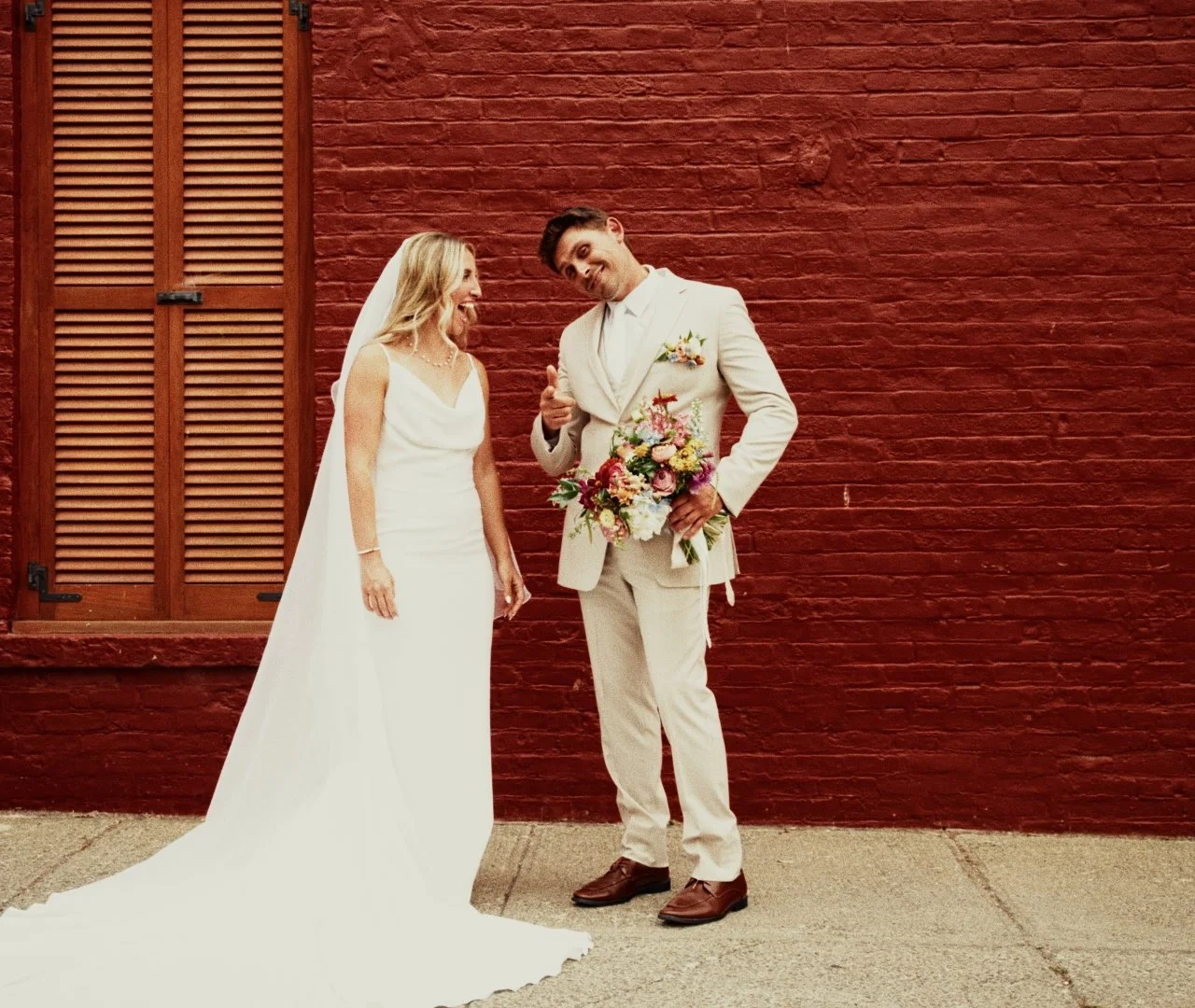 A bride in a white wedding gown and veil, and a groom in a cream suit holding a bouquet, standing in front of a red brick wall, smiling and looking at each other.