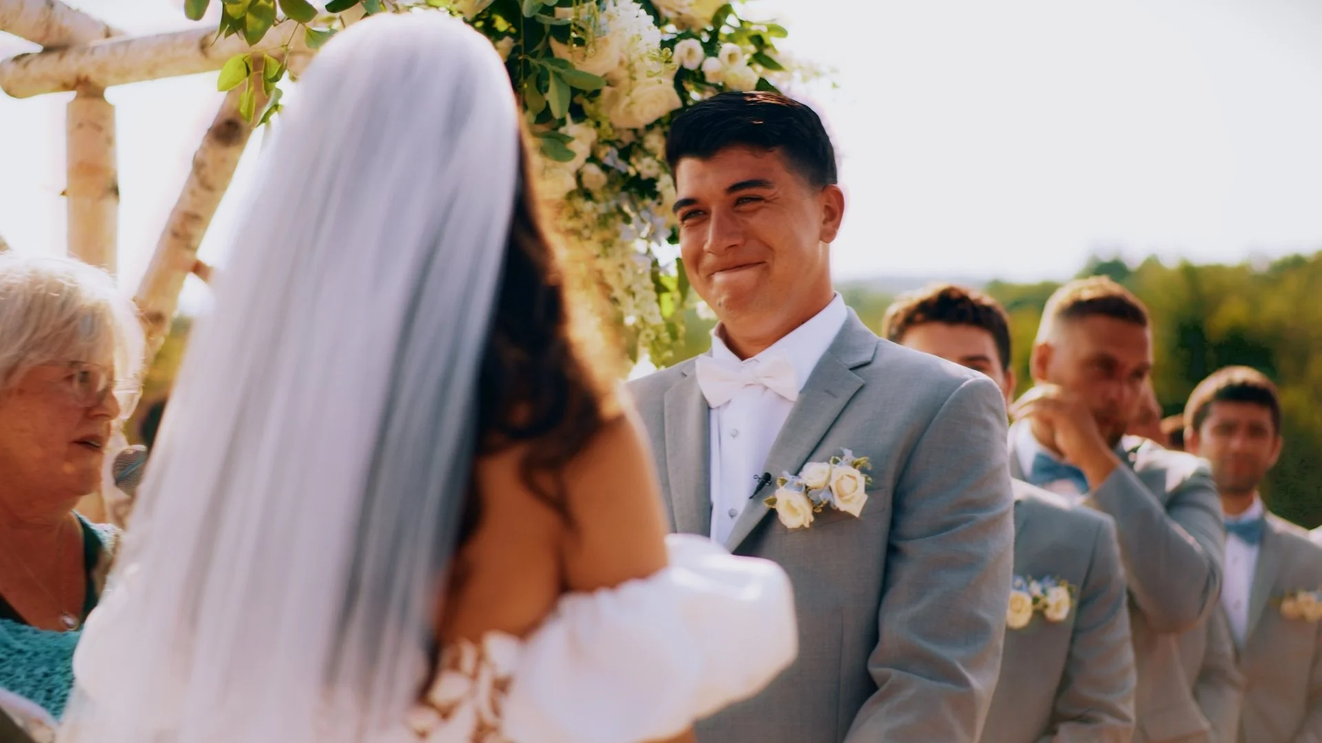 A wedding ceremony outdoors with the groom smiling at the bride, who is wearing a white dress and veil. In the background, groomsmen are seen standing under an arch decorated with flowers, and a woman with glasses is speaking into a microphone.
