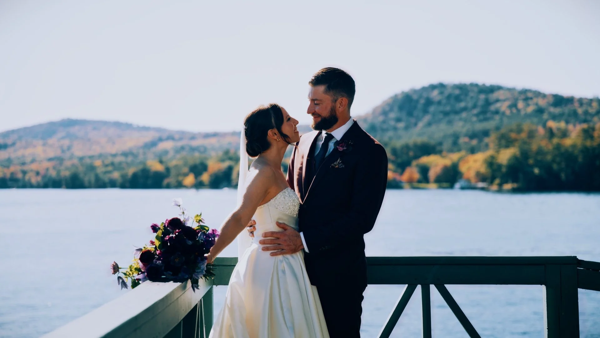 A bride and groom standing close together on a dock by a lake, with mountains and trees in the background, during their wedding celebration.