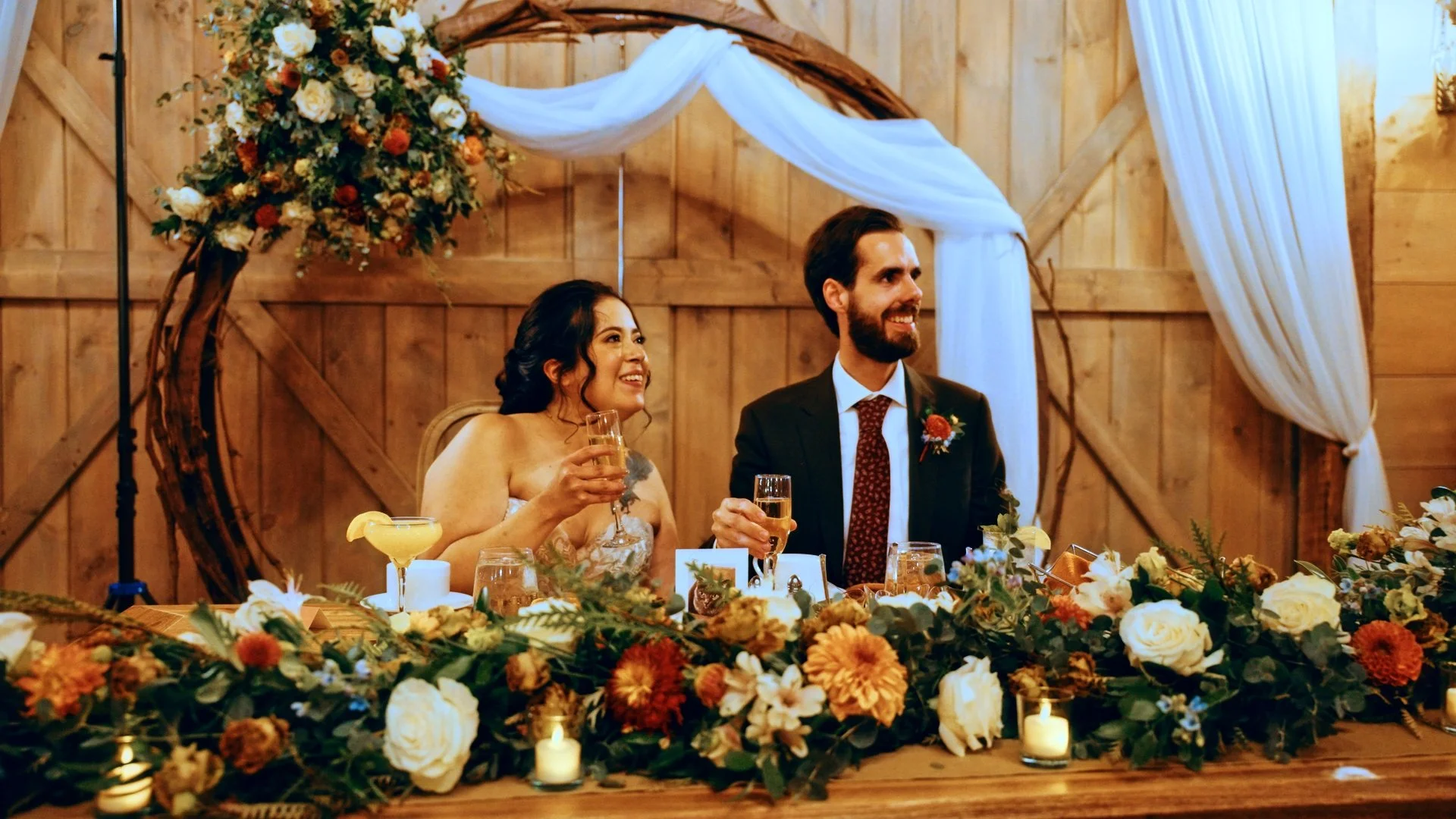 A bride and groom sitting at a wedding reception table decorated with flowers and candles, holding glasses of champagne, in a rustic wooden venue.