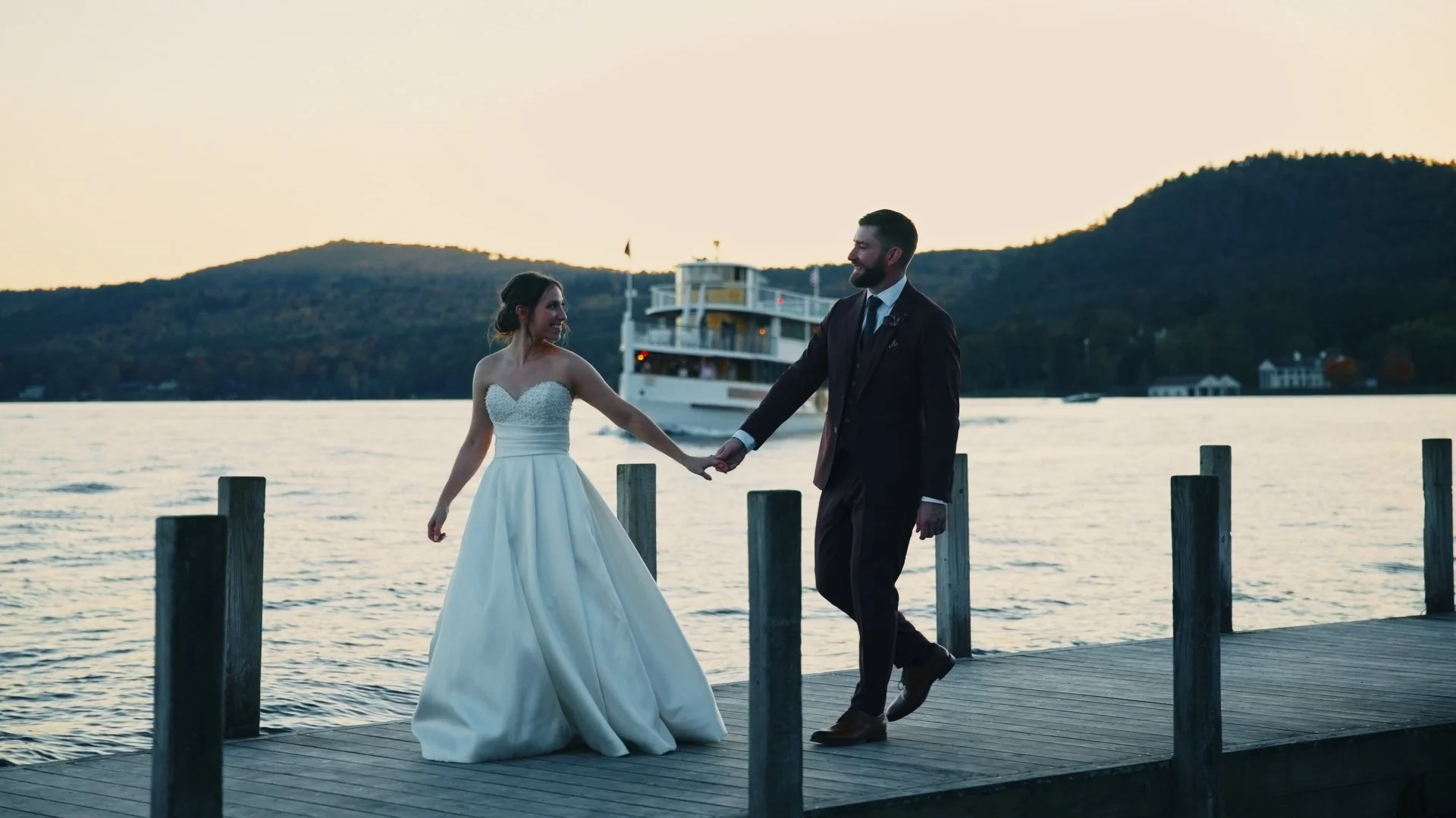 A newlywed couple holding hands and dancing on a wooden dock by a lake at sunset. The bride wears a strapless white wedding gown, and the groom wears a dark suit. A boat is passing by in the background with hills and houses along the shoreline.