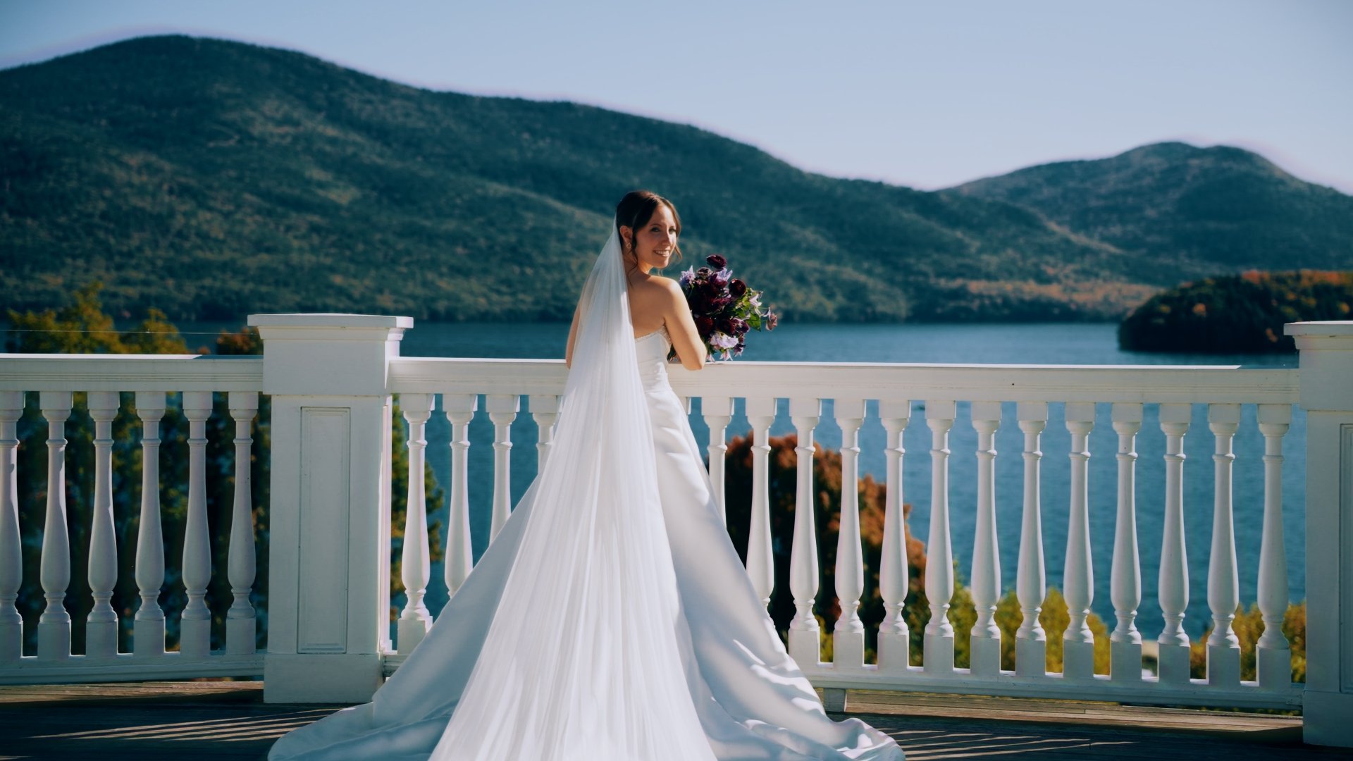 A bride in a white wedding dress and veil holding a bouquet, standing on a balcony with a lake and mountains in the background.