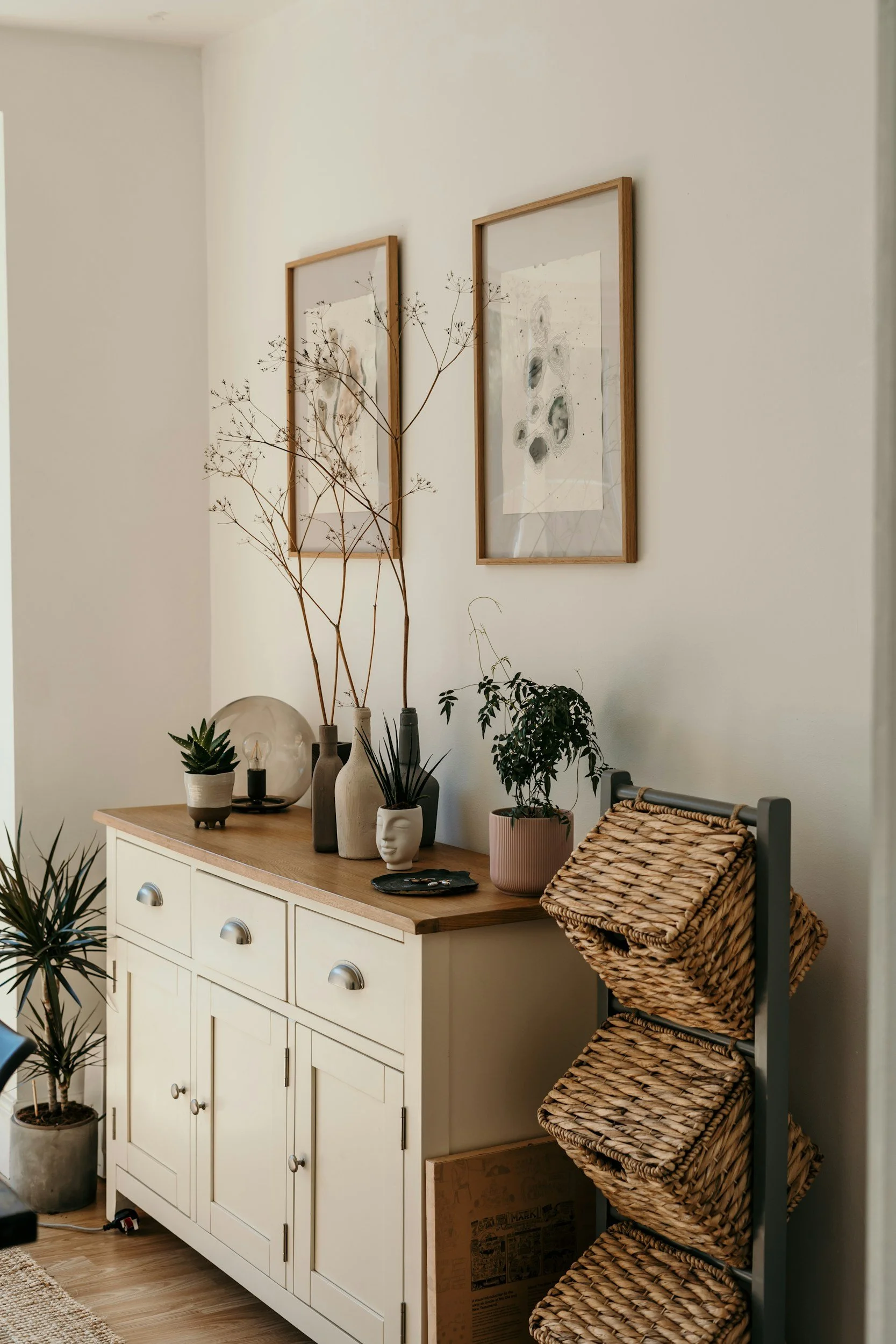 A cream-colored sideboard with three drawers and three cabinet doors, topped with a wooden surface, decorated with various potted plants, vases, and a glass lamp. Above it are two framed abstract art prints on the white wall. To the right, there is a four-tiered woven storage rack with flat woven baskets.
