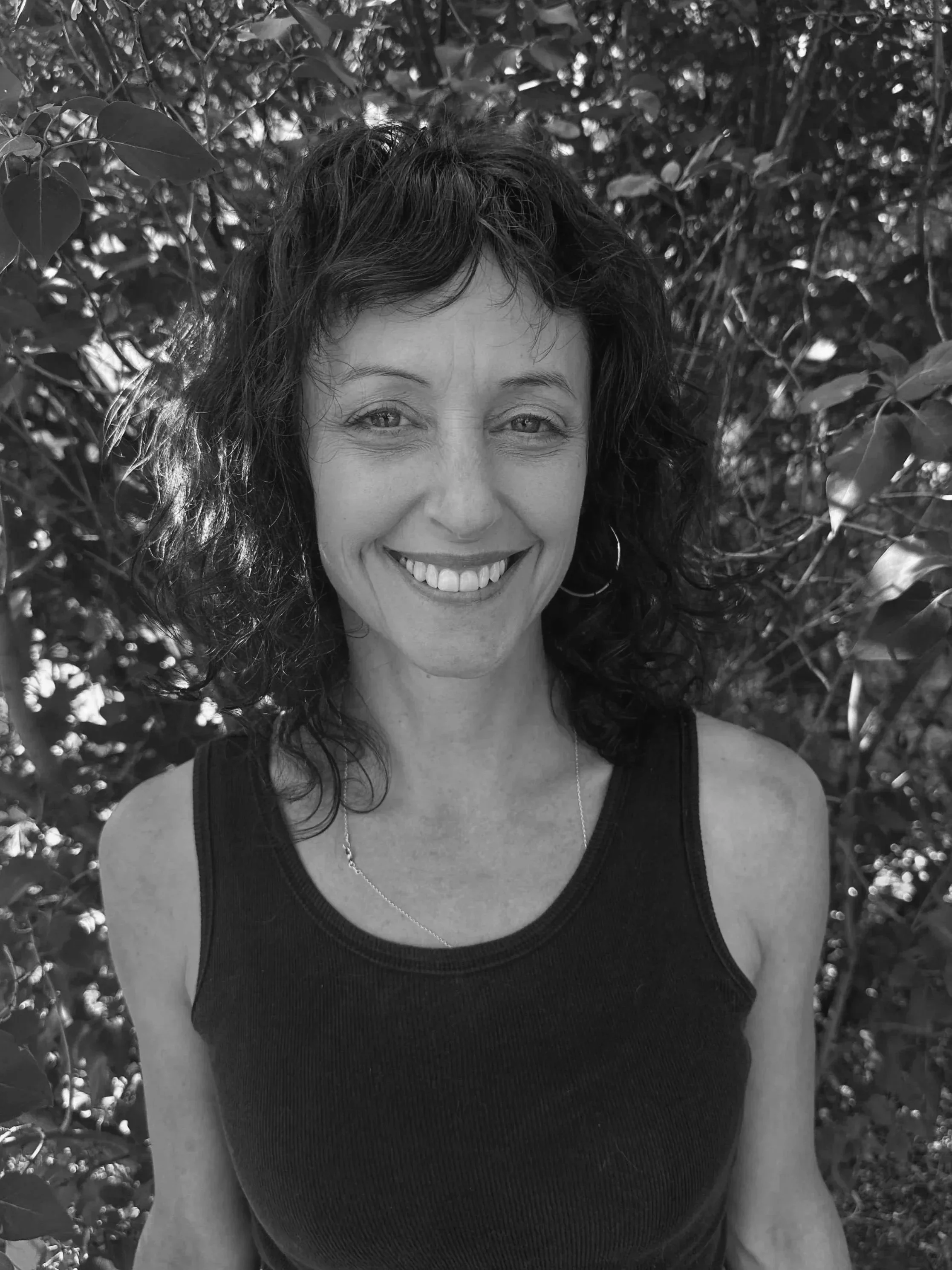 A woman with curly hair smiling outdoors in front of bushes.