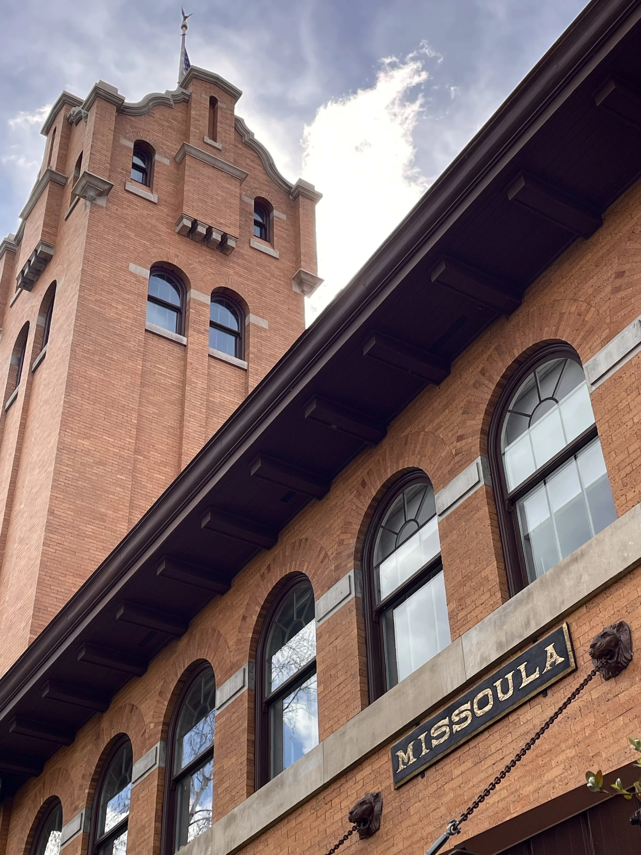 A historic red brick building with arched windows and a tower, featuring a street sign that reads 'MISSOU LA' and decorative lion head fixtures. The sky is partly cloudy with sunlight visible.
