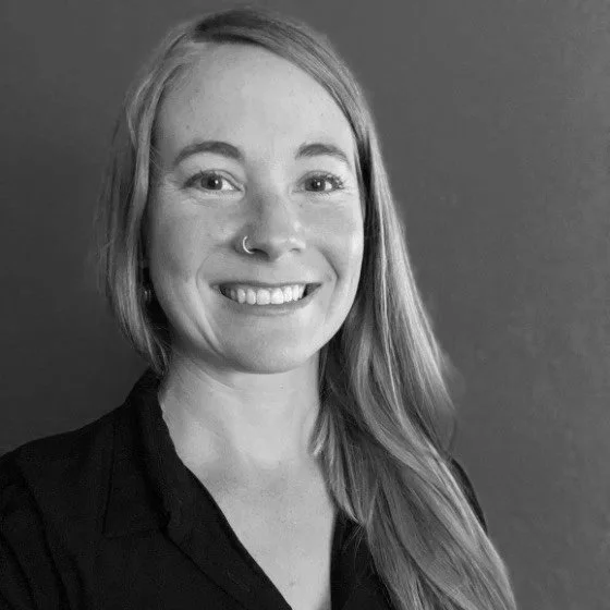 Black and white portrait of a woman with long hair, a nose ring, and a friendly smile, wearing a dark top, standing against a plain background.