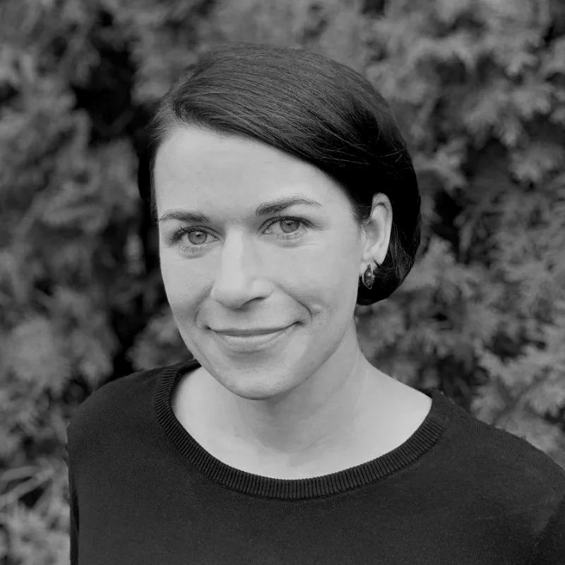 A woman with short dark hair and earrings smiling at the camera, standing outdoors with blurred foliage in the background.