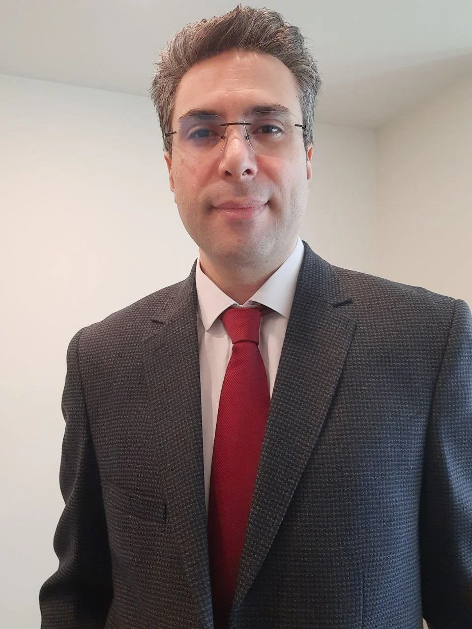 A man wearing glasses, a suit, and a tie taking a selfie in a plain light-colored room.