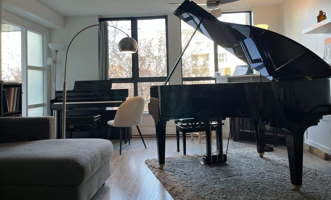 A room with two pianos, one upright and one grand, near large windows with curtains, a beige upholstered chair, a tall floor lamp, a grey rug, and some shelves with books and decorative items.