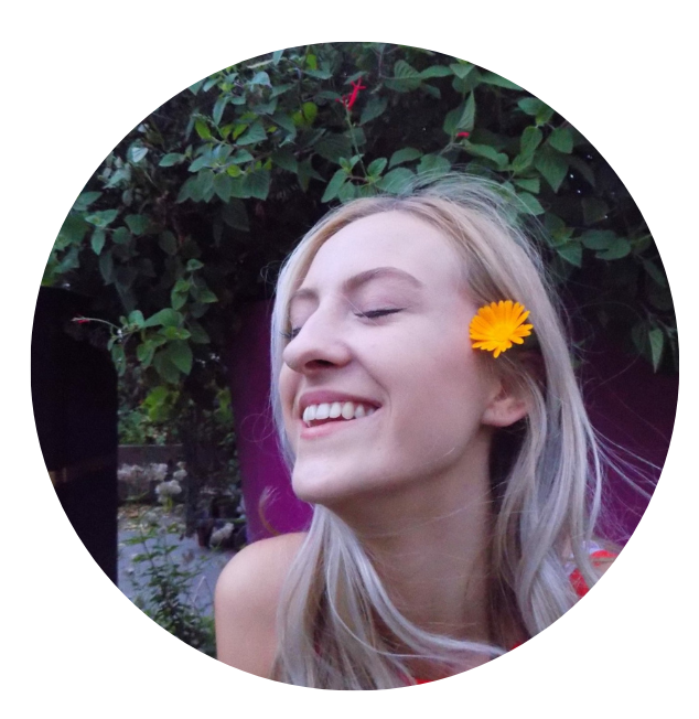 Young woman with long blond hair and a yellow flower behind her ear, smiling with her eyes closed outdoors among green foliage.