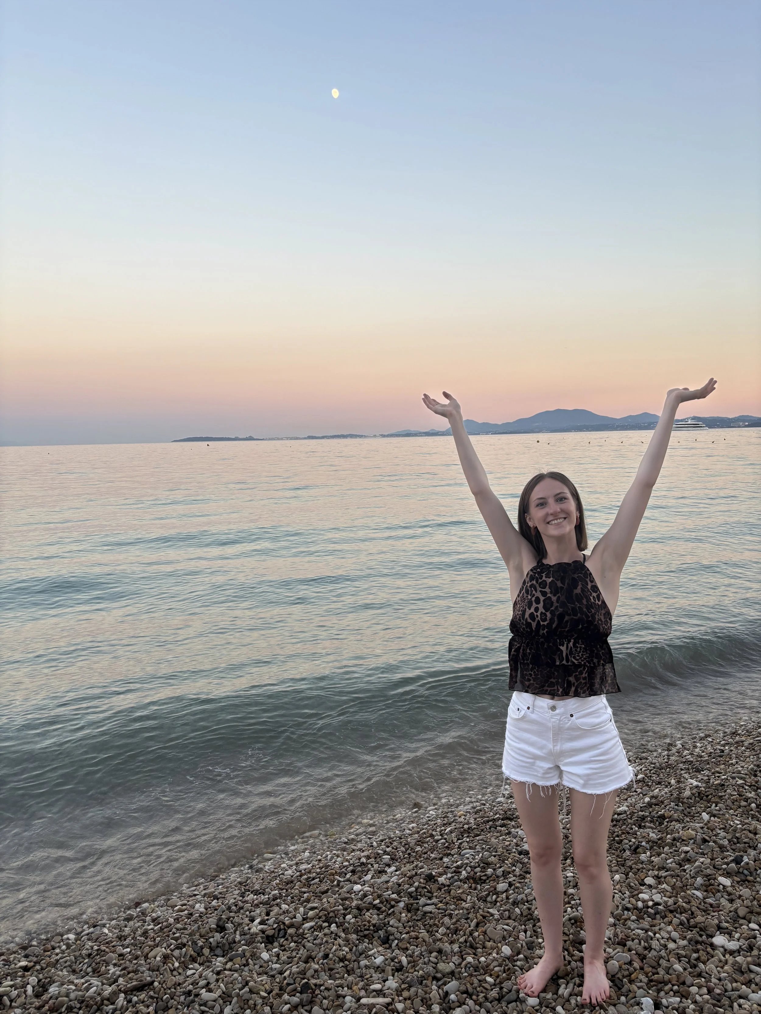 A young woman standing on a rocky beach with water and distant mountains in the background, smiling with arms raised in a 'V' shape, wearing a leopard print sleeveless top and white shorts, during sunset.