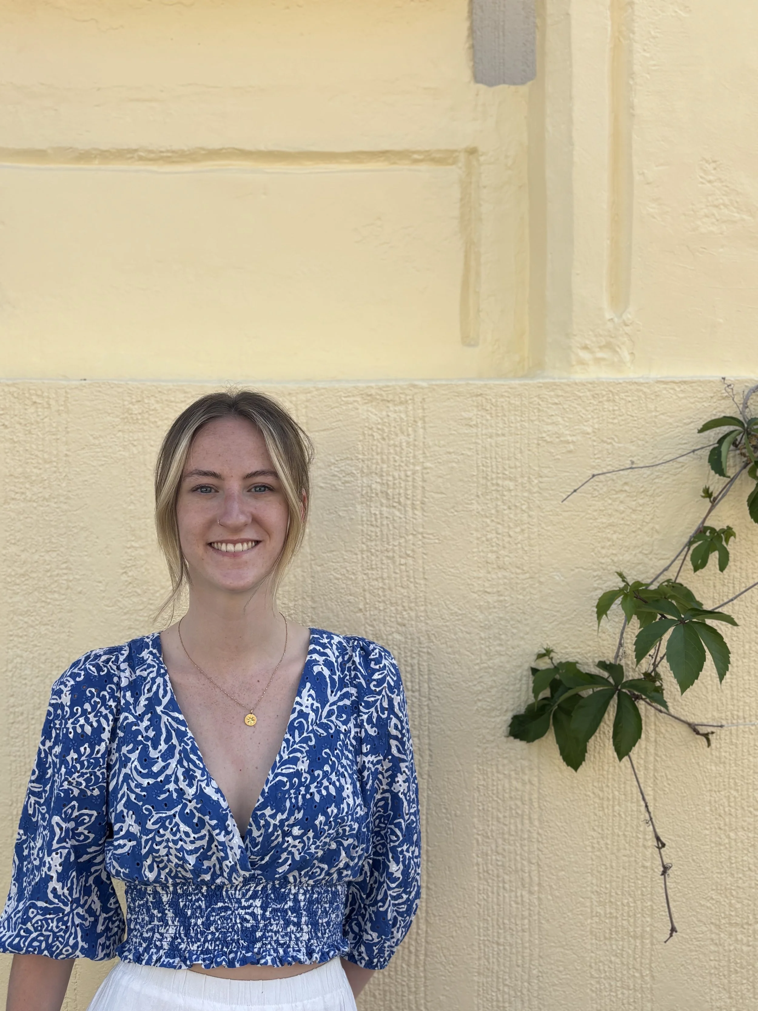 A young woman with blonde hair and a light skin tone smiling, wearing a blue patterned blouse and a gold necklace with a small round pendant, standing in front of a beige textured wall with a green leafy vine on the right.