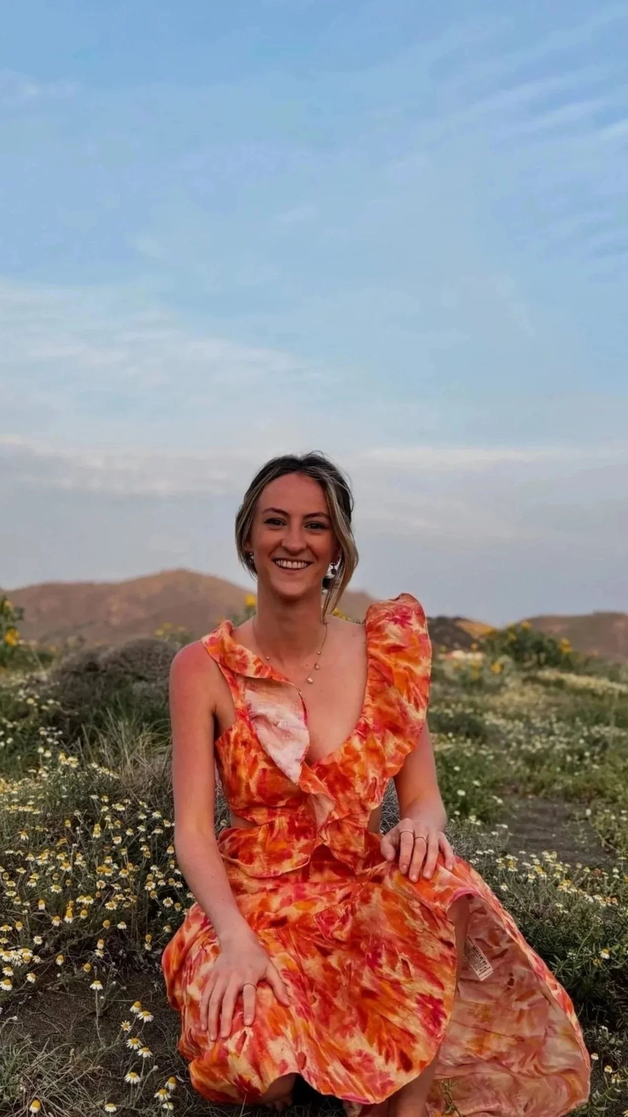 A smiling woman in an orange floral dress sitting outdoors in a field of wildflowers, with hills and a blue sky in the background.