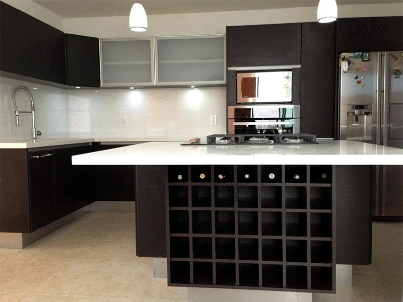 Cocina moderna con gabinetes oscuros, una isla con porta botellas de vino, superficies blancas, estufa empotrada, horno y refrigerador de acero inoxidable.