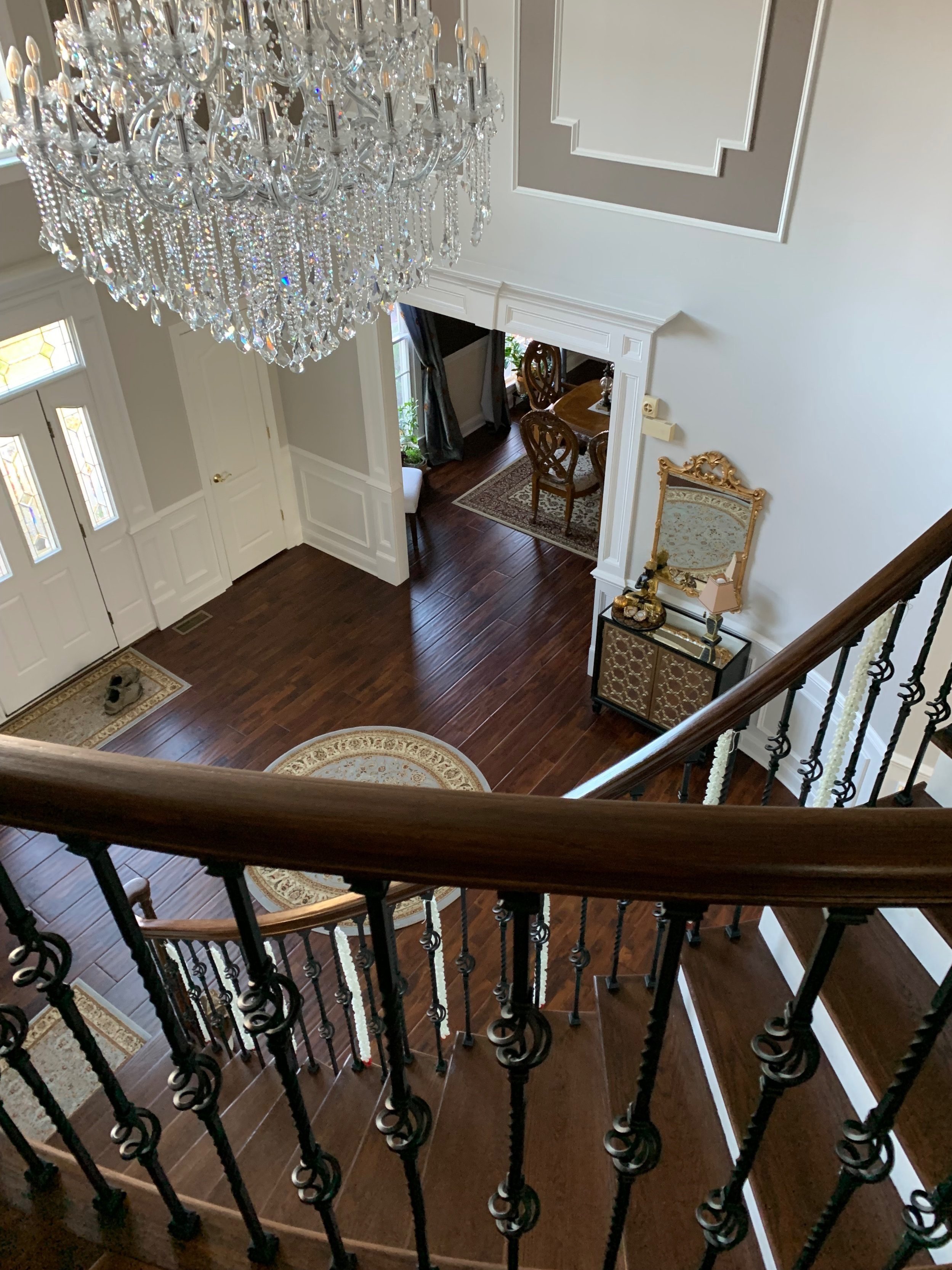 View from a staircase overlooking a spacious, elegant foyer with a chandelier, hardwood floors, a mat by the front door, and a glimpse into a dining area with antique-style chairs and a table.