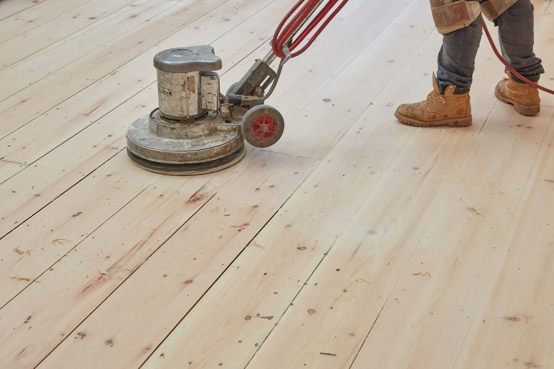 Worker in jeans and work boots sanding a wooden floor with a large industrial floor sander.