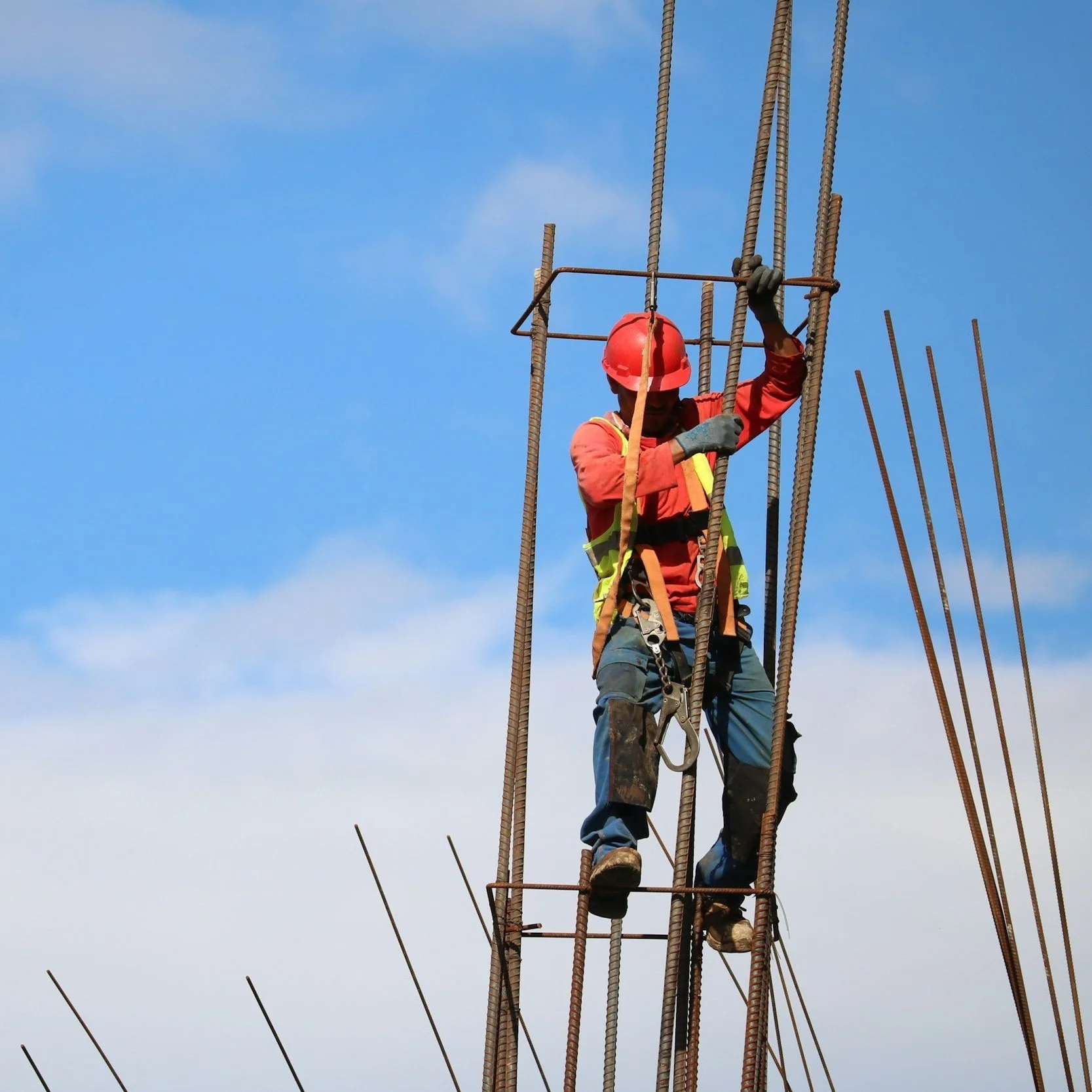 A construction worker in safety gear, including a red helmet and harness, climbing steel rebar at a construction site under a blue sky.