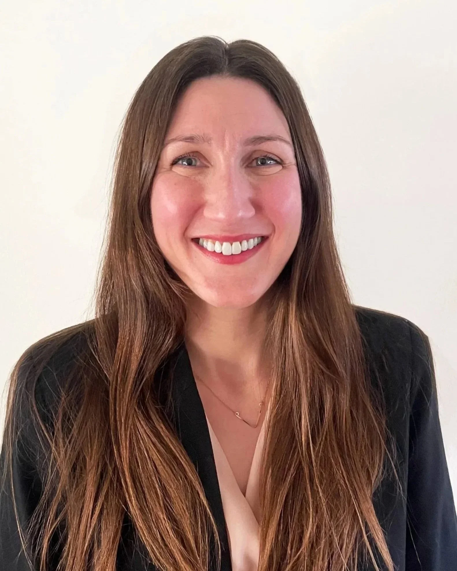A woman smiling with long brown hair, wearing a black blazer and a light pink top, against a plain white background.