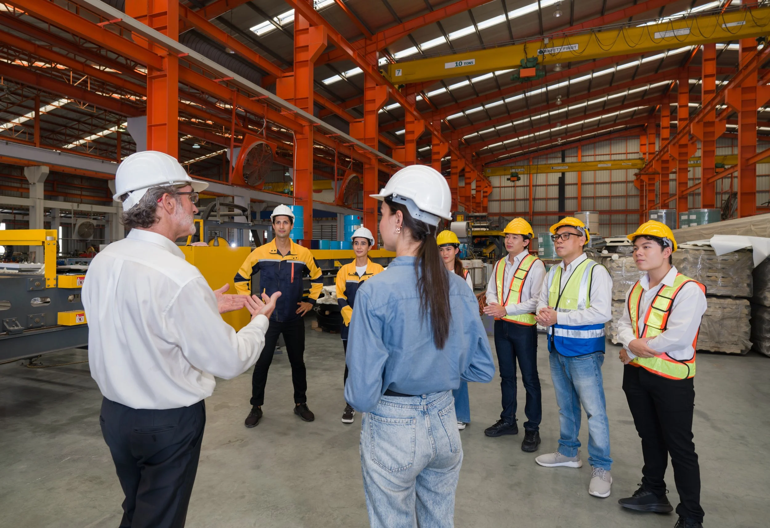 A group of workers with safety helmets in a factory listening to a man in a white shirt explaining something.