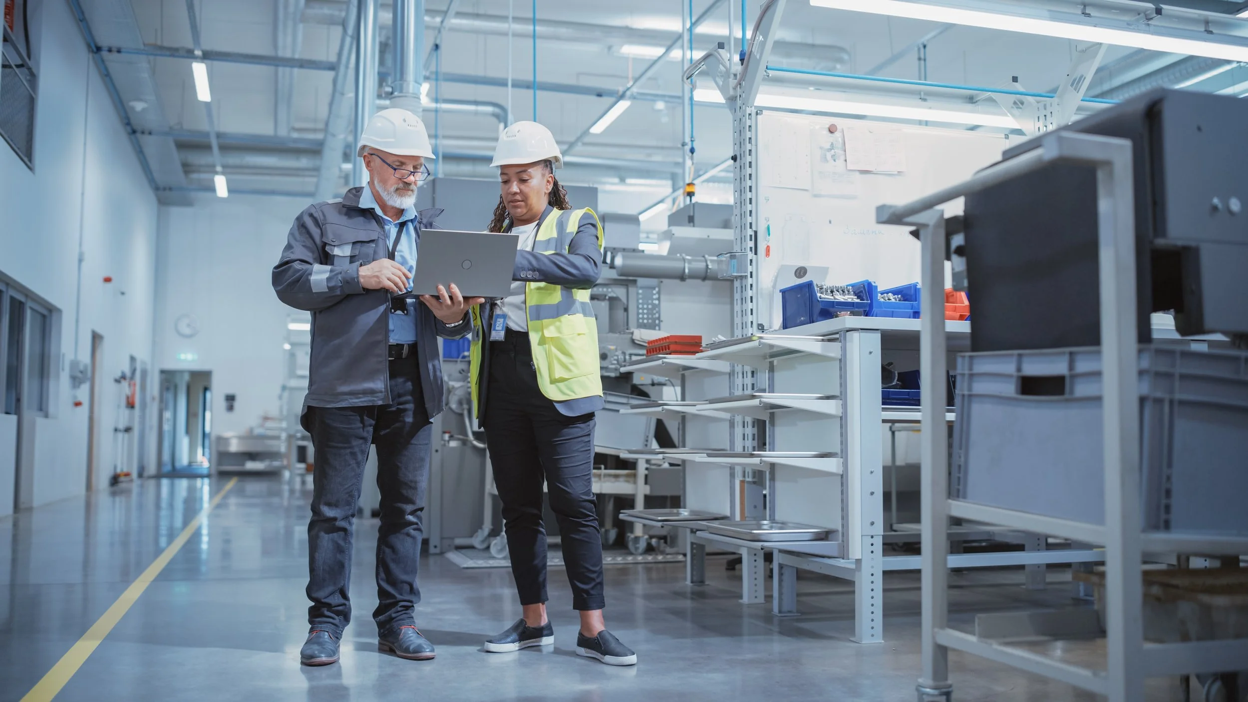 Two engineers in a manufacturing plant wearing hard hats and safety vests looking at a laptop together, surrounded by shelves with parts and tools.