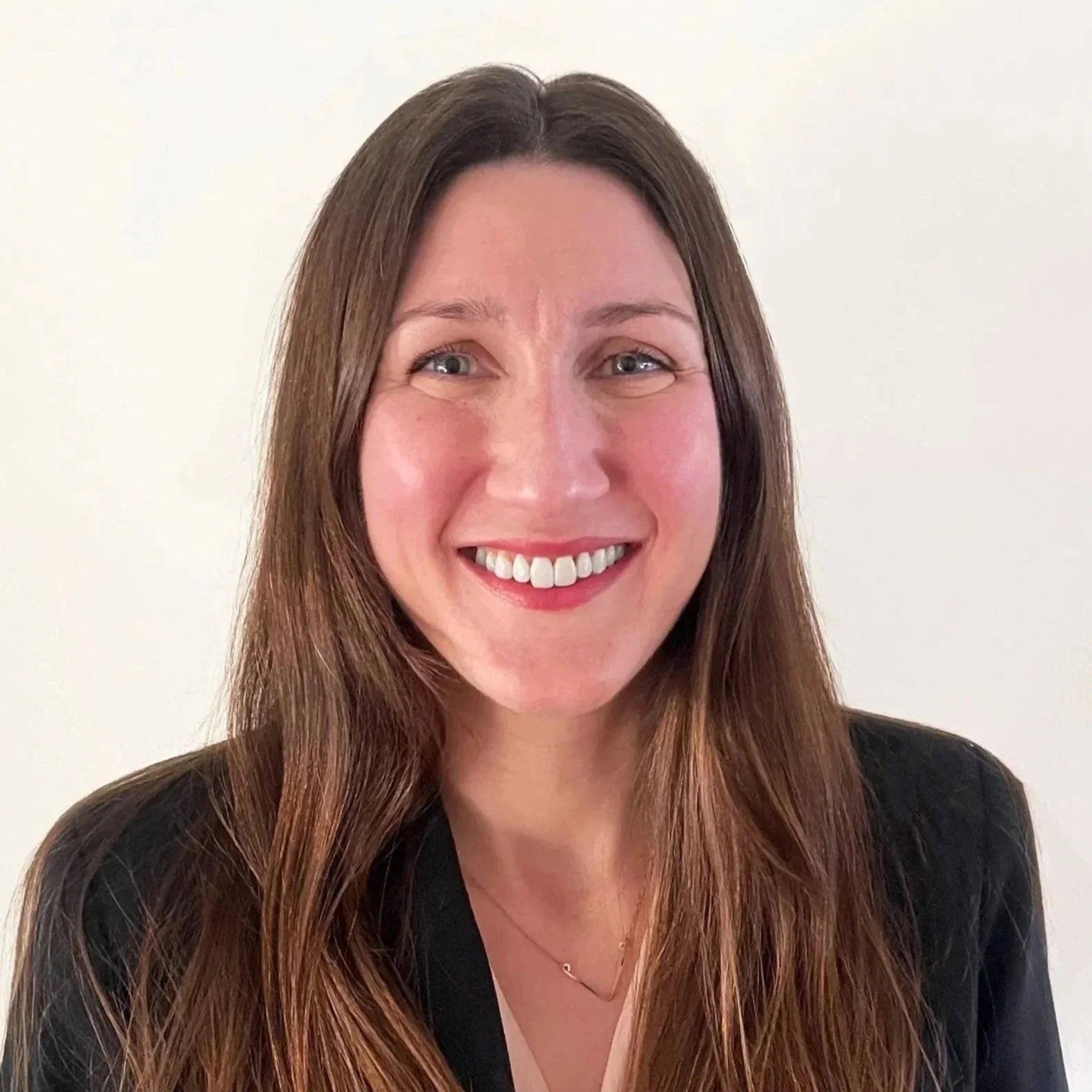 A woman with long brown hair, smiling with teeth visible, wearing a black blazer and a gold necklace.