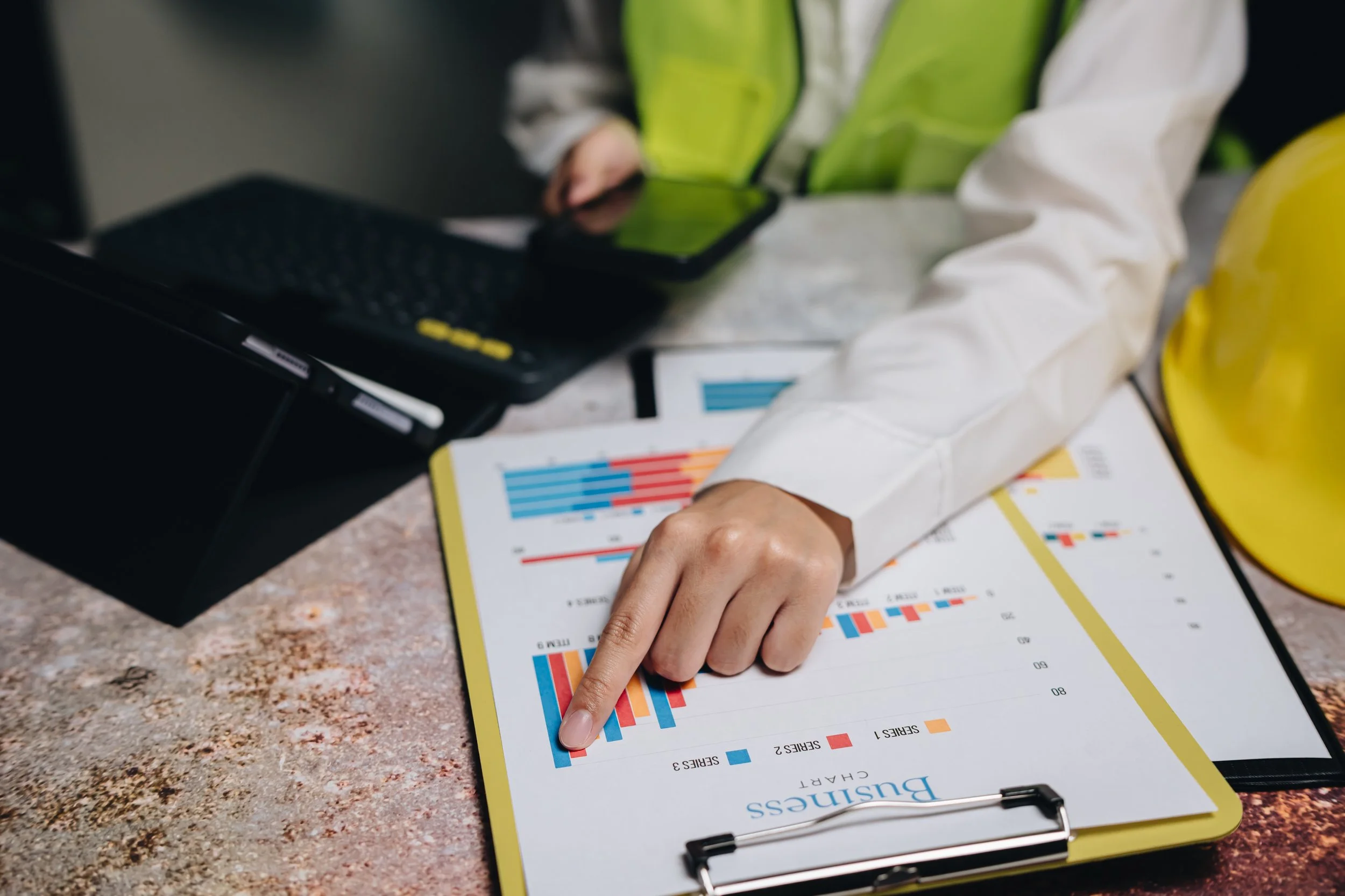Person pointing at bar chart on clipboard with a computer and hard hat nearby.