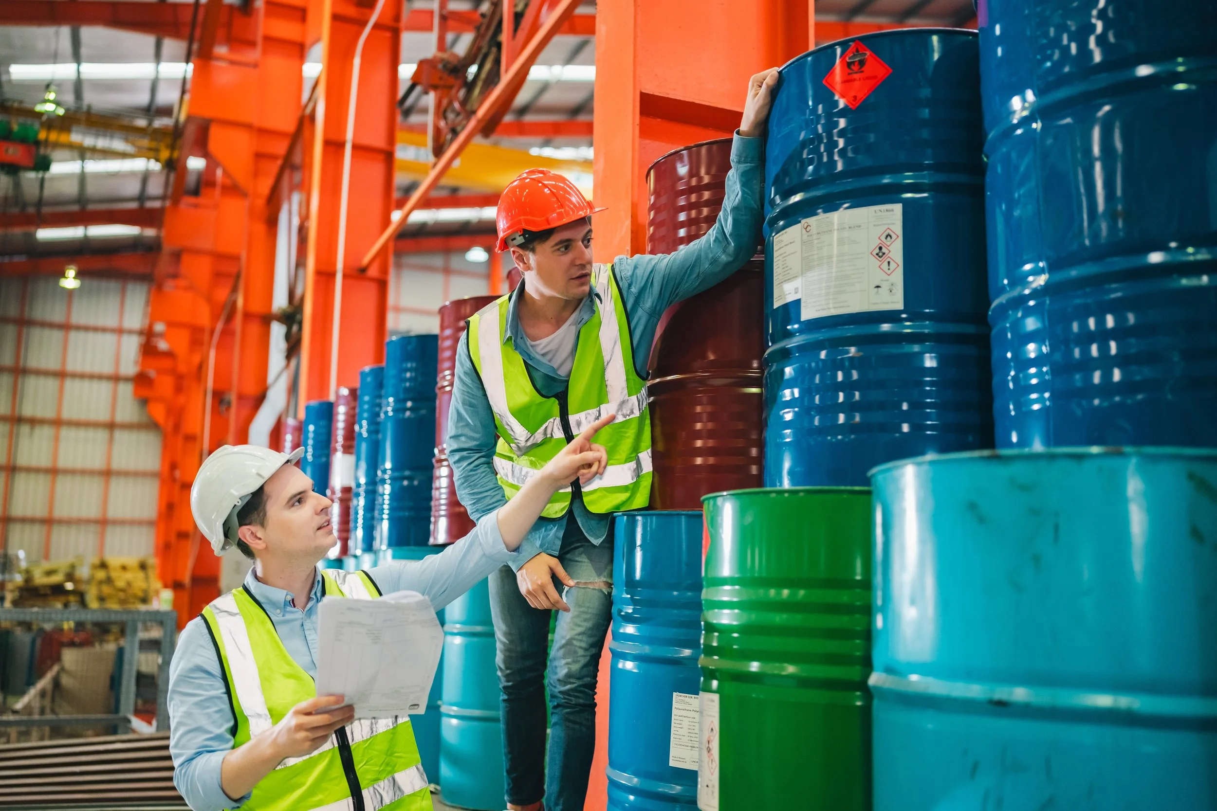 Two workers, a man and a woman, wearing safety helmets and high-visibility vests, inspecting large blue, red, and green industrial drums in a warehouse.