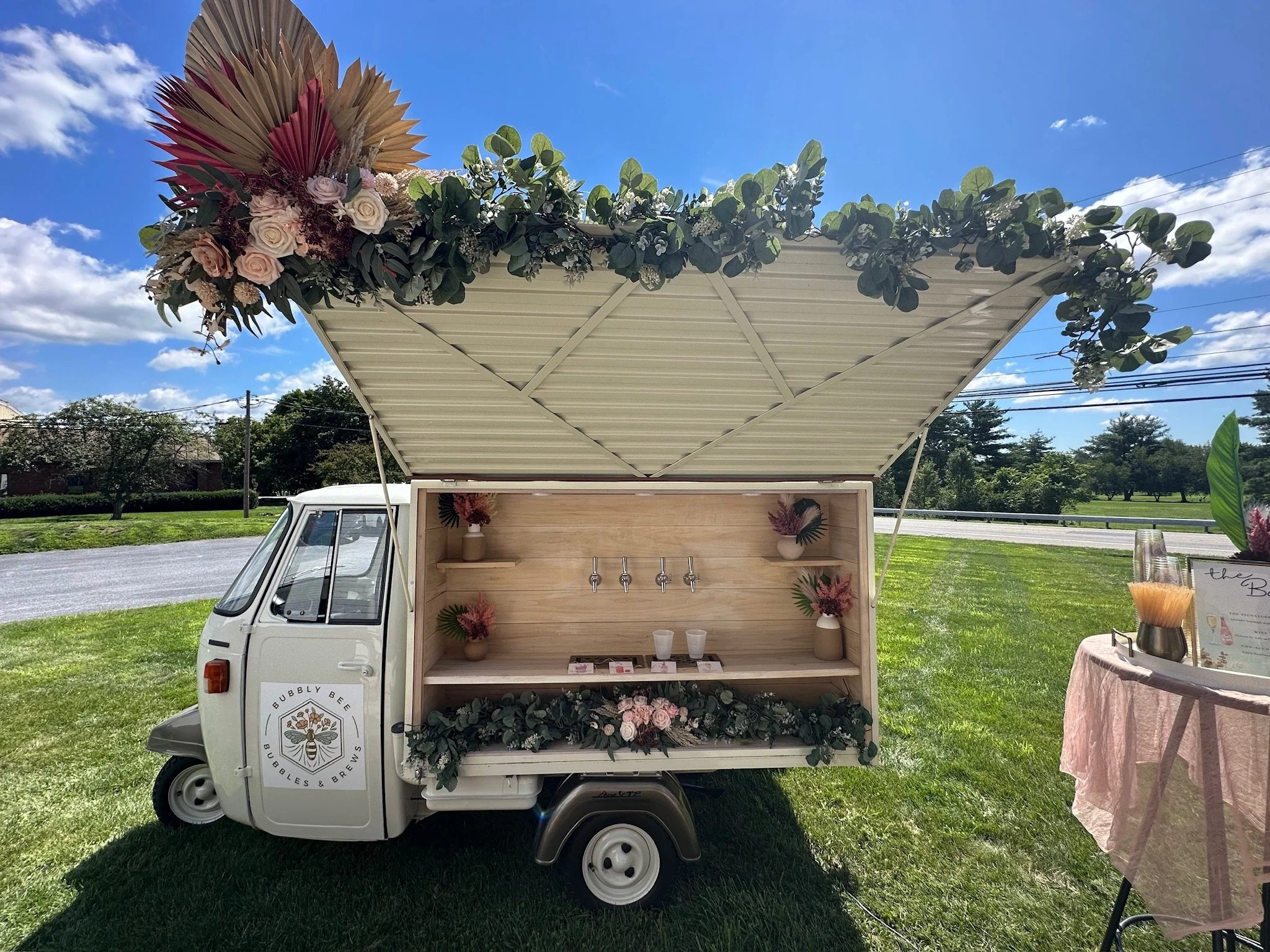 A mini mobile bar decorated with floral arrangements, including roses and greenery, under a large open canopy in an outdoor setting on a sunny day.